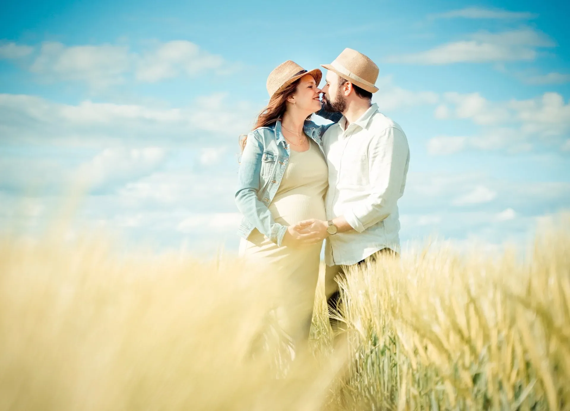 Babybauch Shooting Outdoor - Ein verliebtes Paar steht in einem Weizenfeld und blickt sich liebevoll an, während sie sich an den Händen halten, bei sonnigem Himmel.