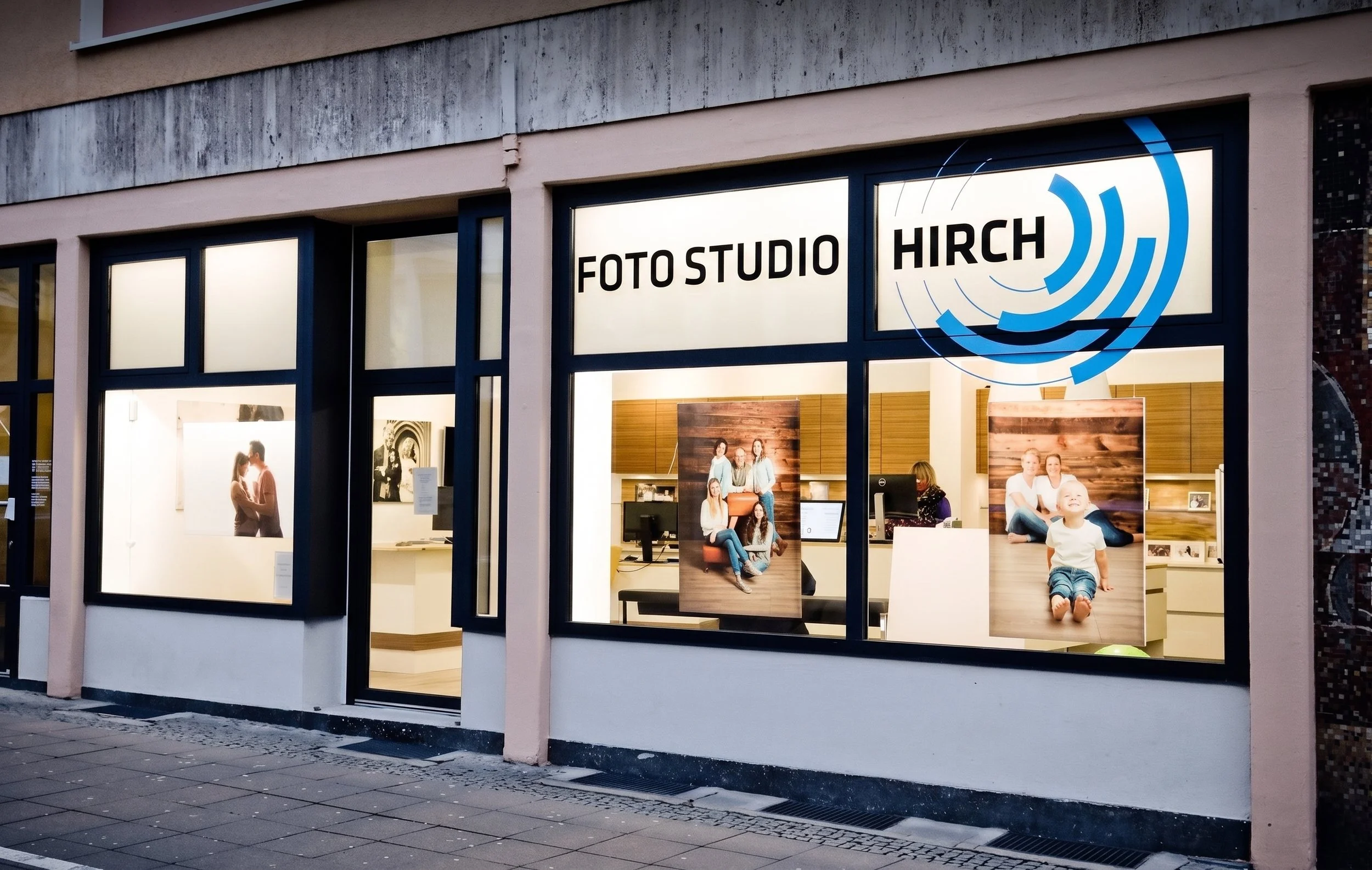 Das Fenster des Fotostudio Hirch in Darmstadt mit schwarzen Rahmen, innen Menschen beim Fotoshooting, ein Poster mit einer Gruppe von jungen Frauen und einem Kind, das auf dem Boden sitzt, ist sichtbar.
