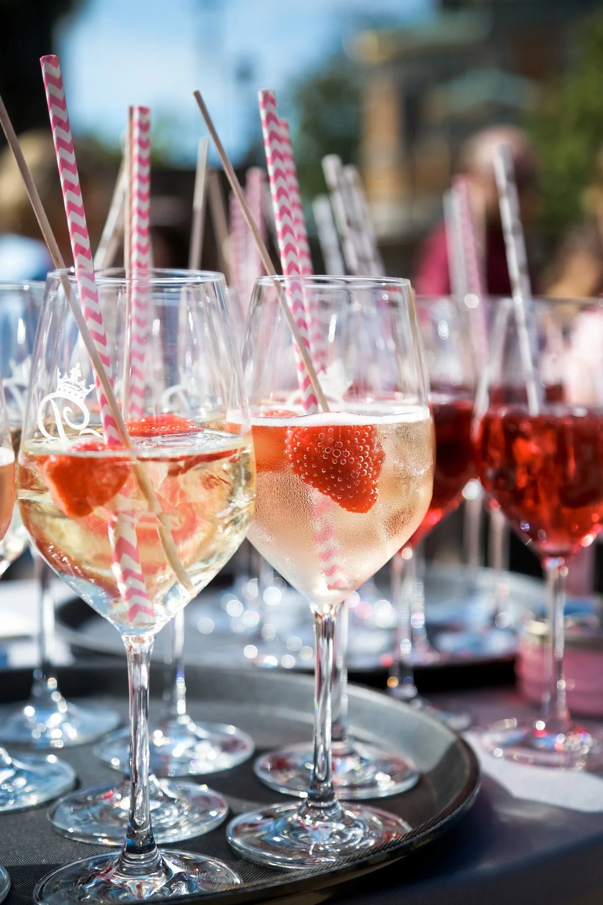 Mehrere Weingläser mit Sekt, die mit Erdbeeren und Strohhalmen gefüllt sind, auf einem schwarzen Tablett, im Hintergrund ist ein unscharfer Garten oder Außenbereich zu sehen.