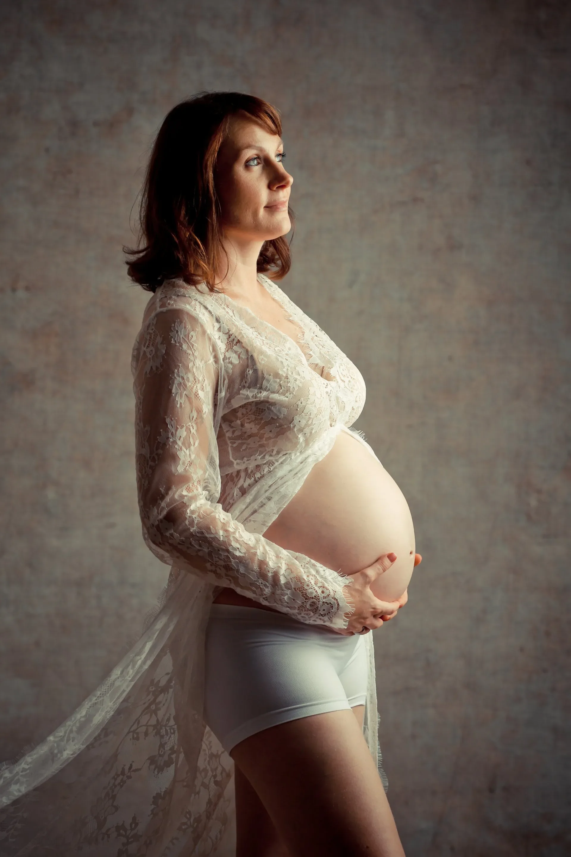 Eine schwangere Frau in einem weißen Spitzenkleid, die seitlich steht und ihre Hände auf ihren Bauch legt, vor einem neutralen Hintergrund.