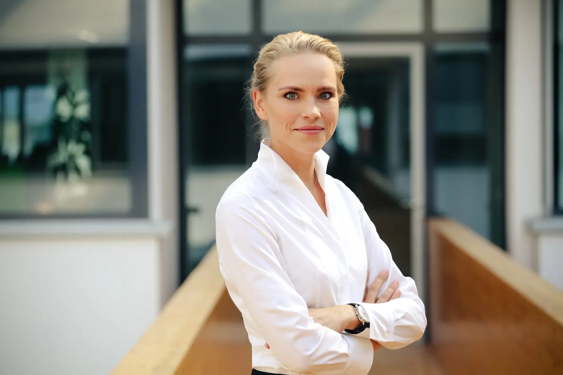 Eine elegante Frau mit blonden Haaren, die einen weißen Hemd trägt, steht im modernen Bürogebäude mit großen Fenstern im Hintergrund.