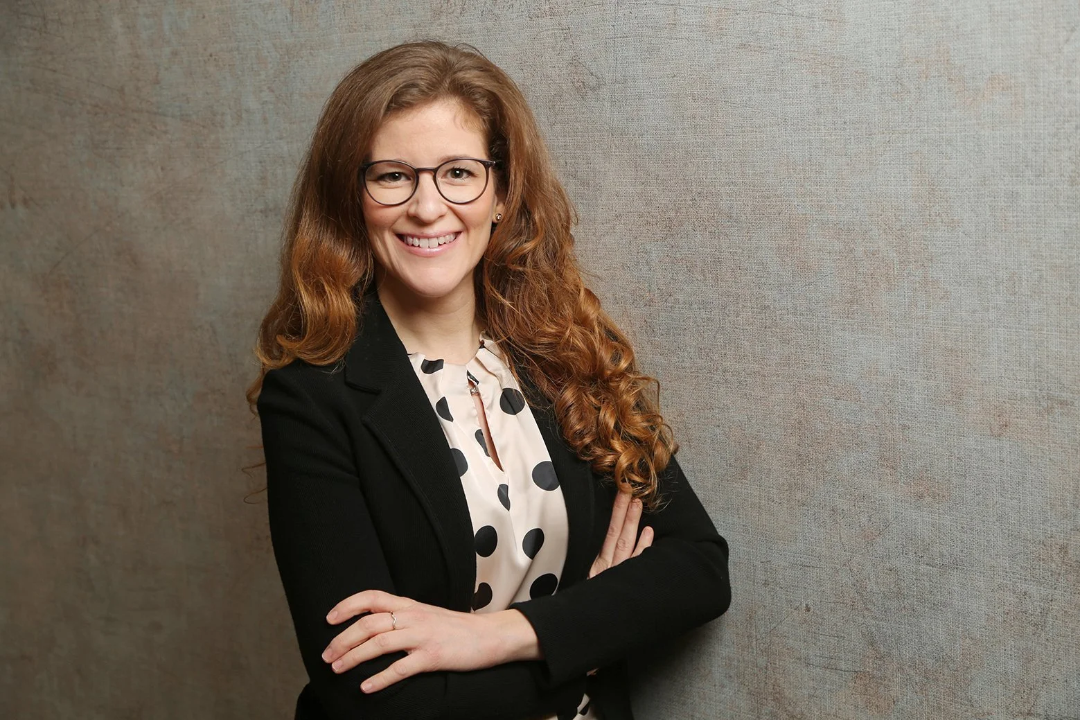 Bewerbungsbild - Eine lächelnde Frau mit roten, lockigen Haaren und Brille, die vor einer grauen, leicht strukturierte Wand steht, getragen in einem schwarzen Blazer und einem weißen Hemd mit schwarzen Pouf.