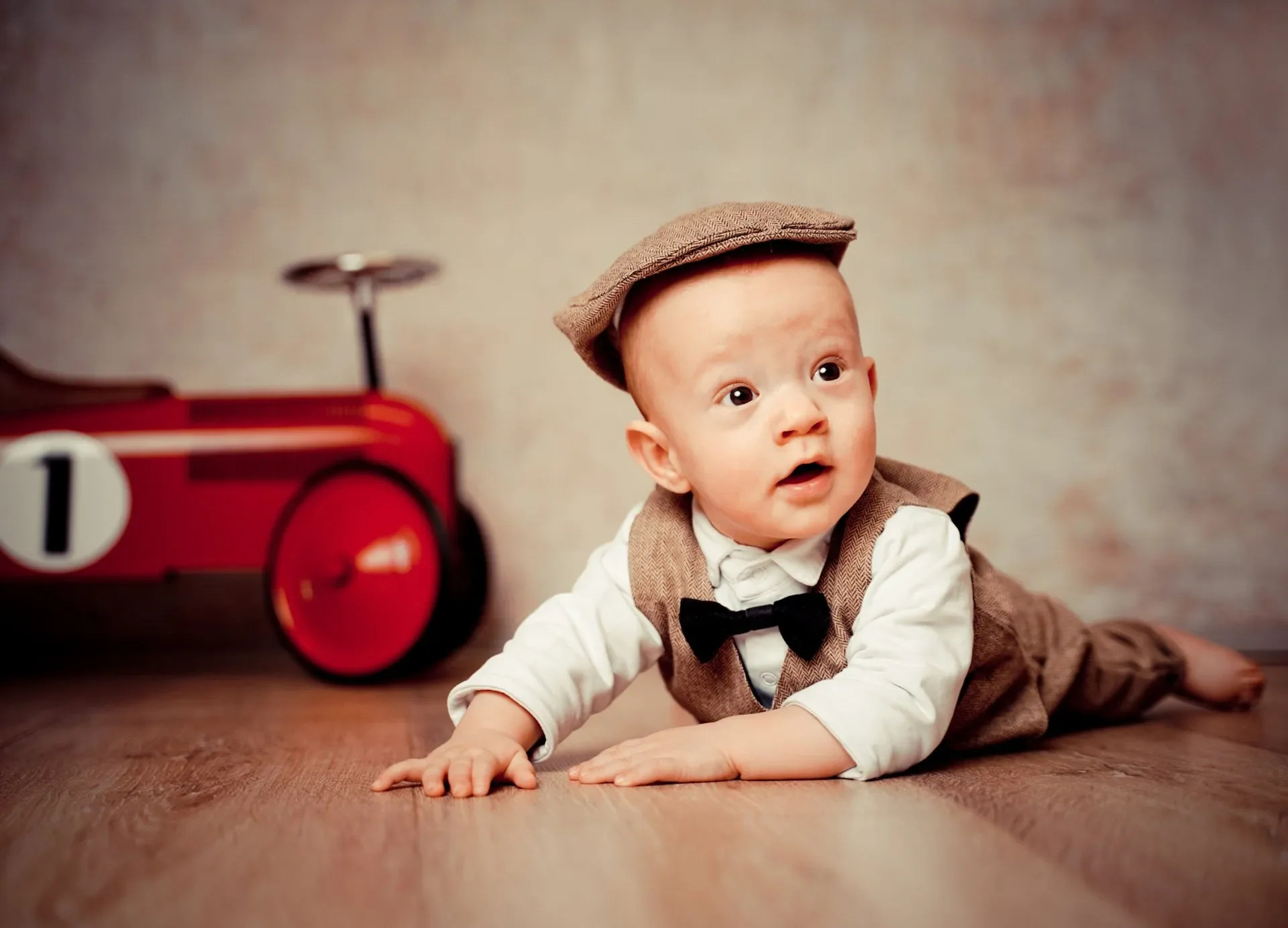 Kleiner Junge trägt einen braunen Tweed-Hut, ein weißes Hemd, einen schwarzen Bow-Tie und eine braune Weste mit Knöpfen, liegt auf dem Boden und schaut neugierig nach vorne. Im Vordergrund ist eine rote Spielzeugkamera sichtbar.