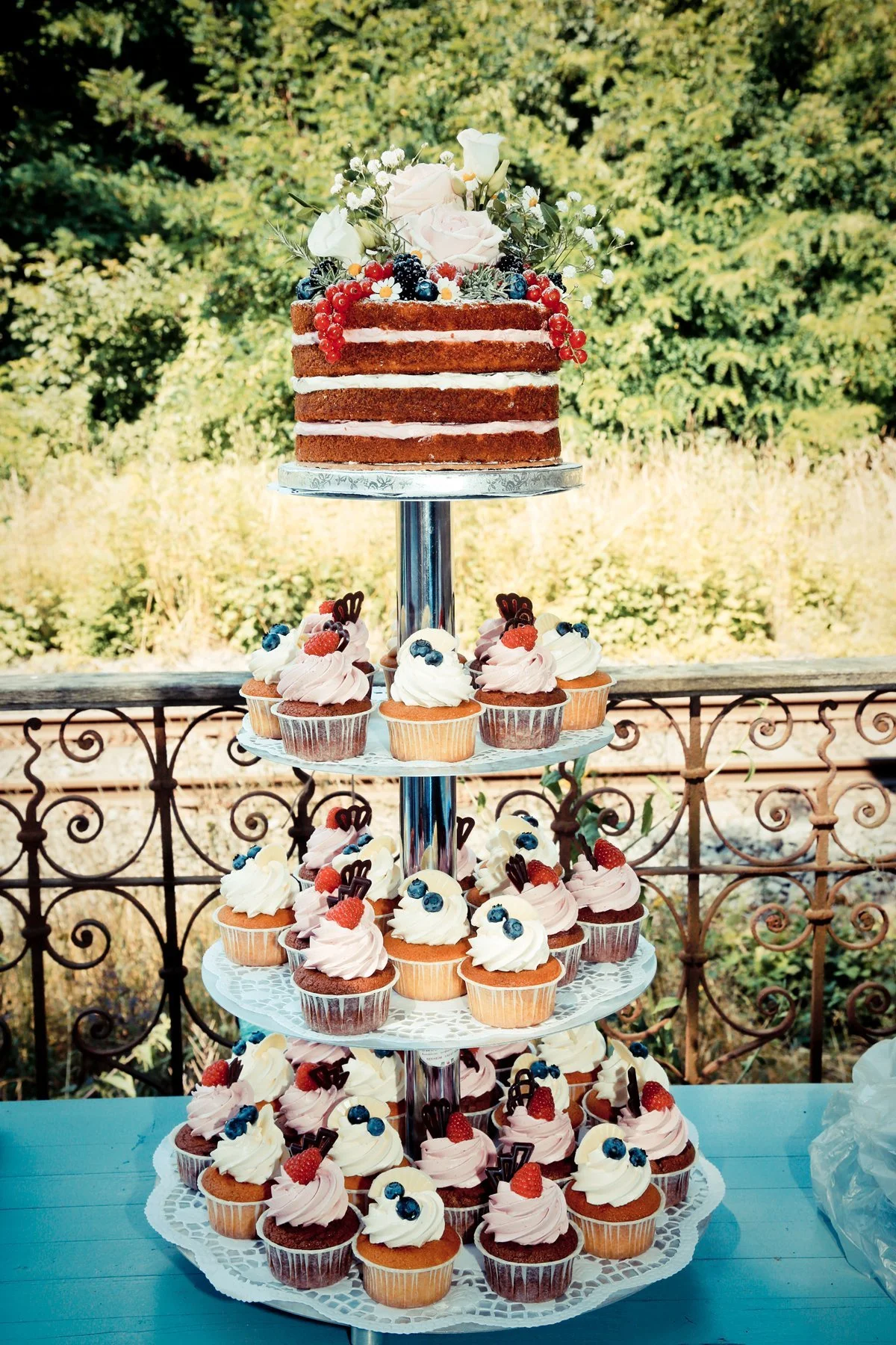 Mehrstöckiger Kuchen mit Frühlingsblumen und Beeren auf der obersten Schicht, umgeben von Cupcakes mit Beeren und Schokoladenstückchen, auf einer Terrasse mit Garten im Hintergrund.