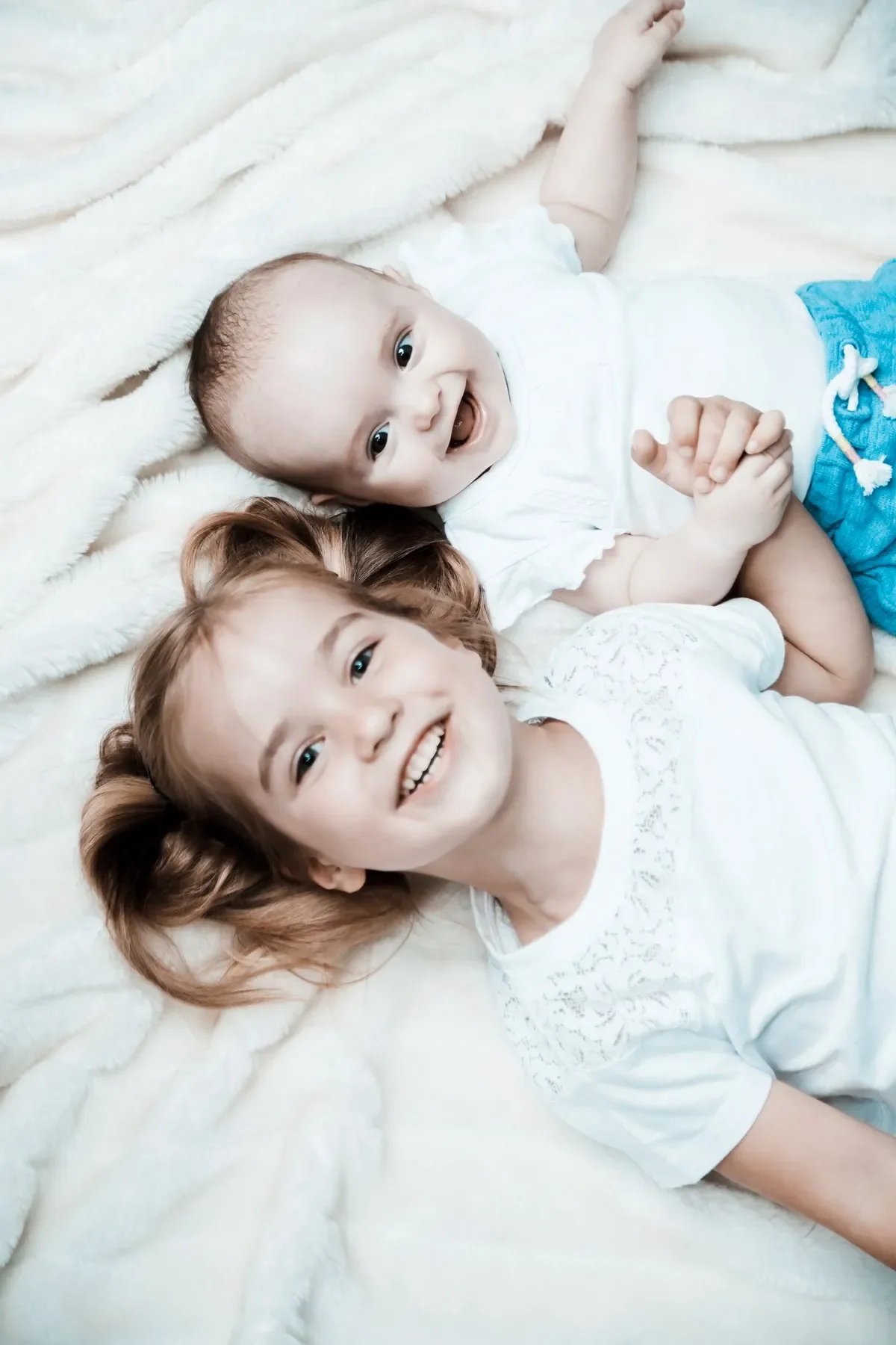 Zwei glückliche Kinder, ein Junge und ein Mädchen, liegen auf einem weißen, kuscheligen Bett und lächeln in die Kamera.