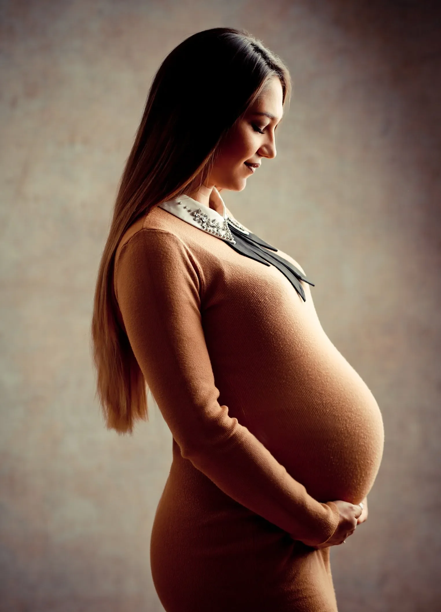 Schwangere Frau in einem braunen Kleid, die ihre Hände auf den Bauch legt, seitlich gesehen, vor neutralem Hintergrund.