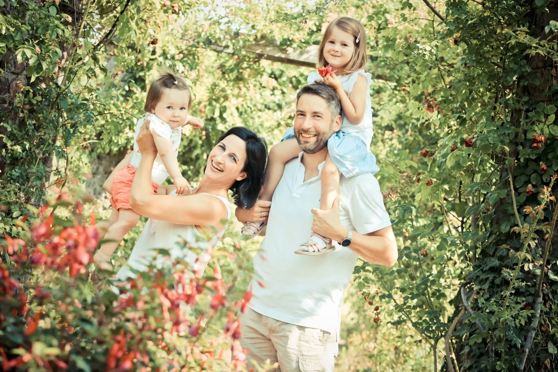 Familie mit zwei kleinen Mädchen im Garten zwischen Rosensträuchern, die im Sommer sitzen und lachen.
