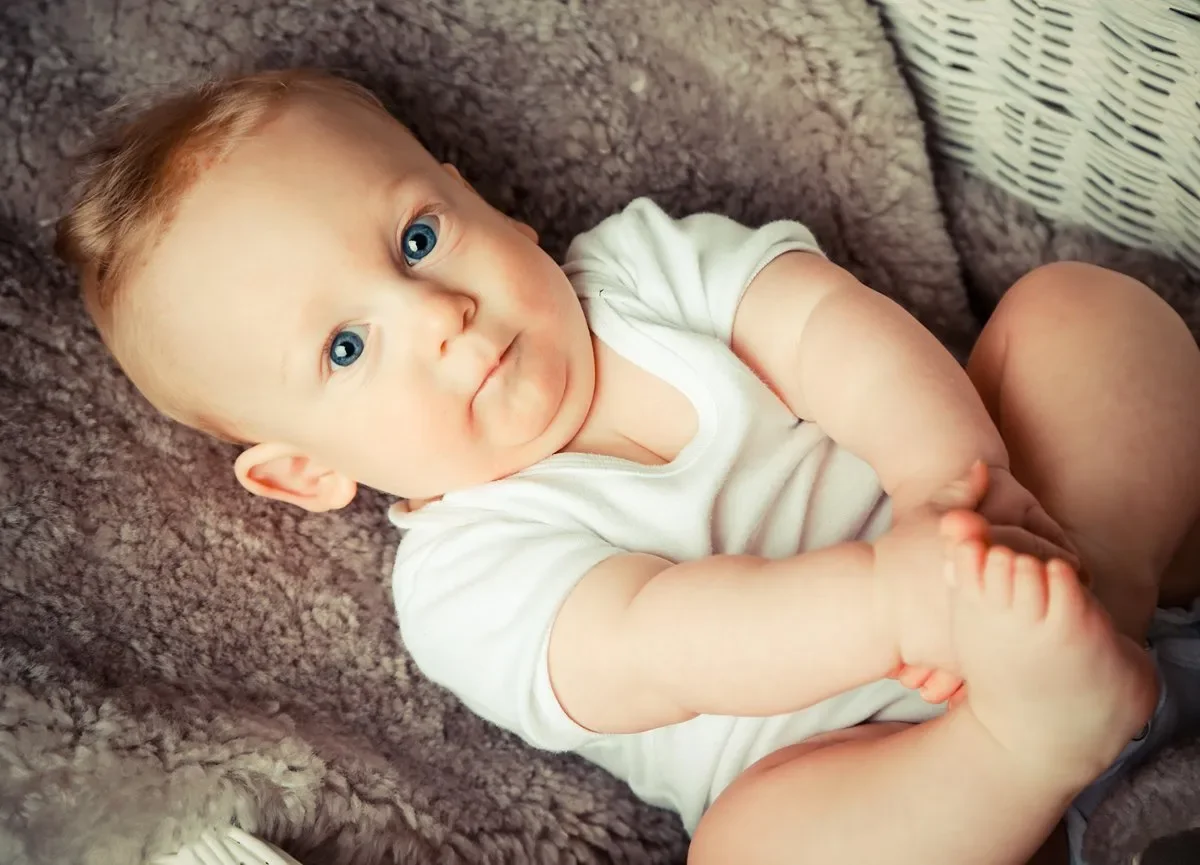 Ein Baby mit blauen Augen liegt auf einem weichen, braunen Teppich, trägt ein weißes T-Shirt und schaut direkt in die Kamera.