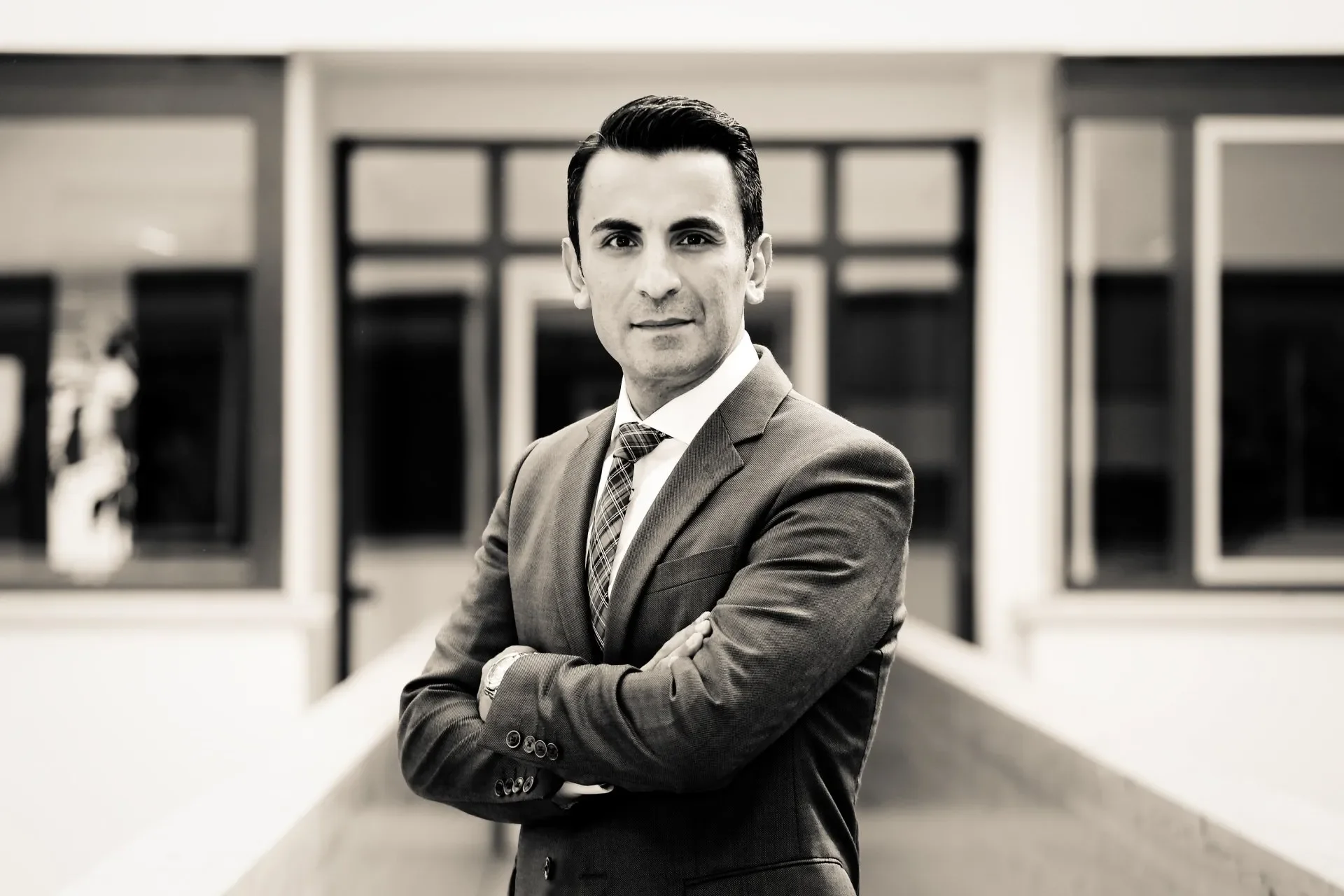 Ein professionell gekleideter Mann mit dunklem, gut frisiertem Haar posiert vor einem modernen Gebäude mit großen Fenstern, in einer schwarzen und weißen Fotografie.