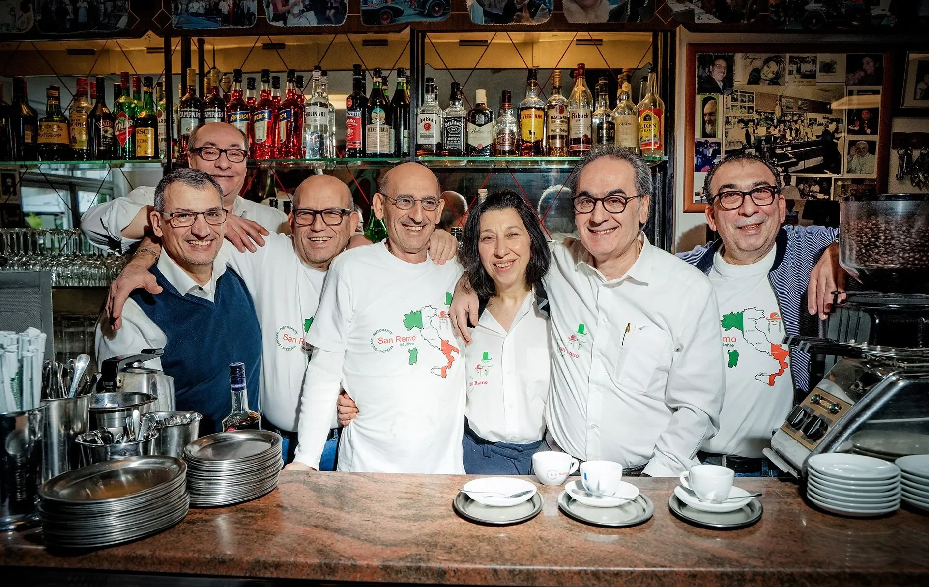 Eine Gruppe von sieben Menschen, die in einer Bar oder einem Restaurant stehen, alle lächeln und sich umarmen. Hinter ihnen sind Regale mit verschiedenen Spirituosen. Im Vordergrund befinden sich Teller, Kaffeetassen und Küchenutensilien auf einer Th