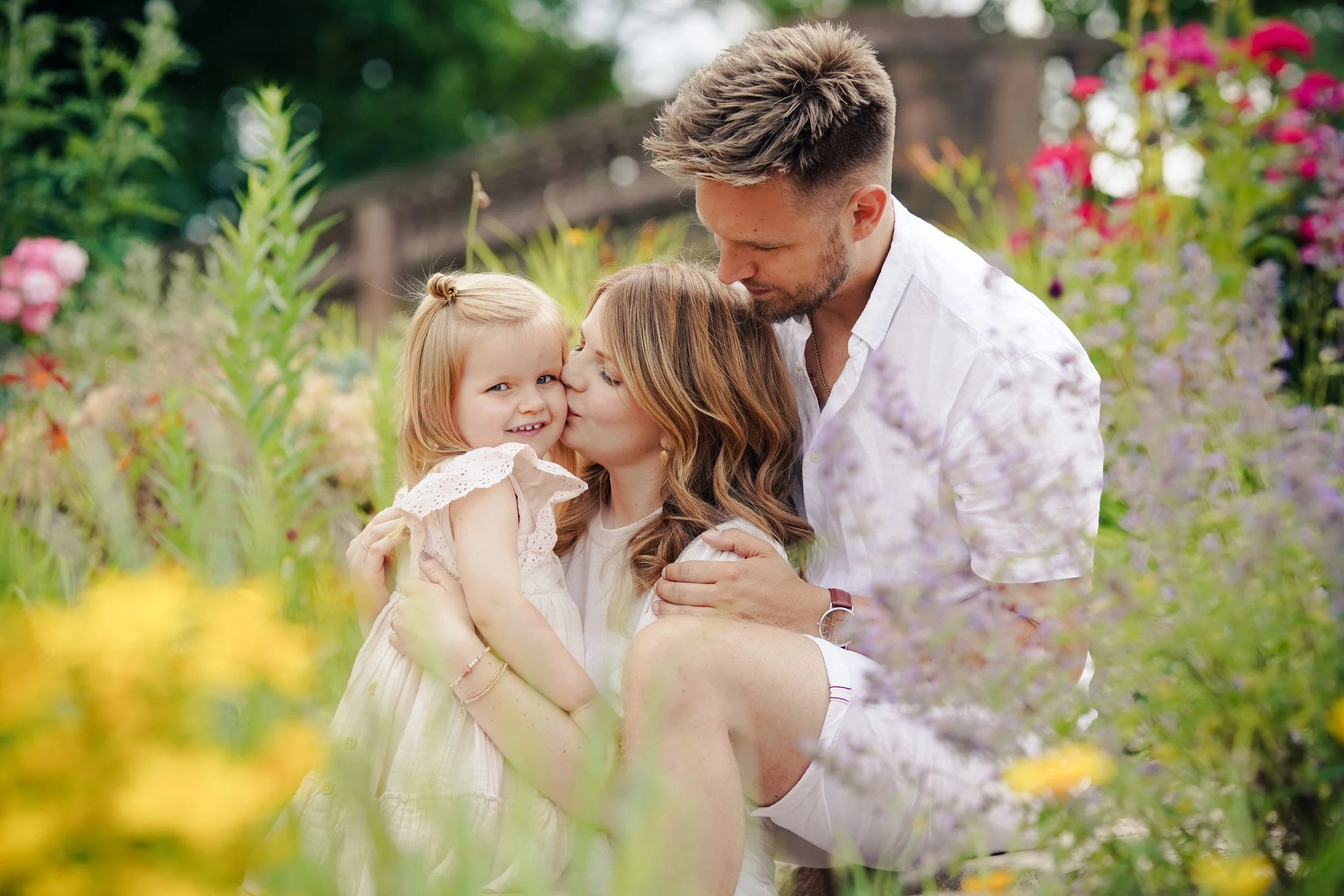 Familie im Garten, Kind wird von Mutter geküsst, Vater hält sie, im Hintergrund Blumen und Bäume.