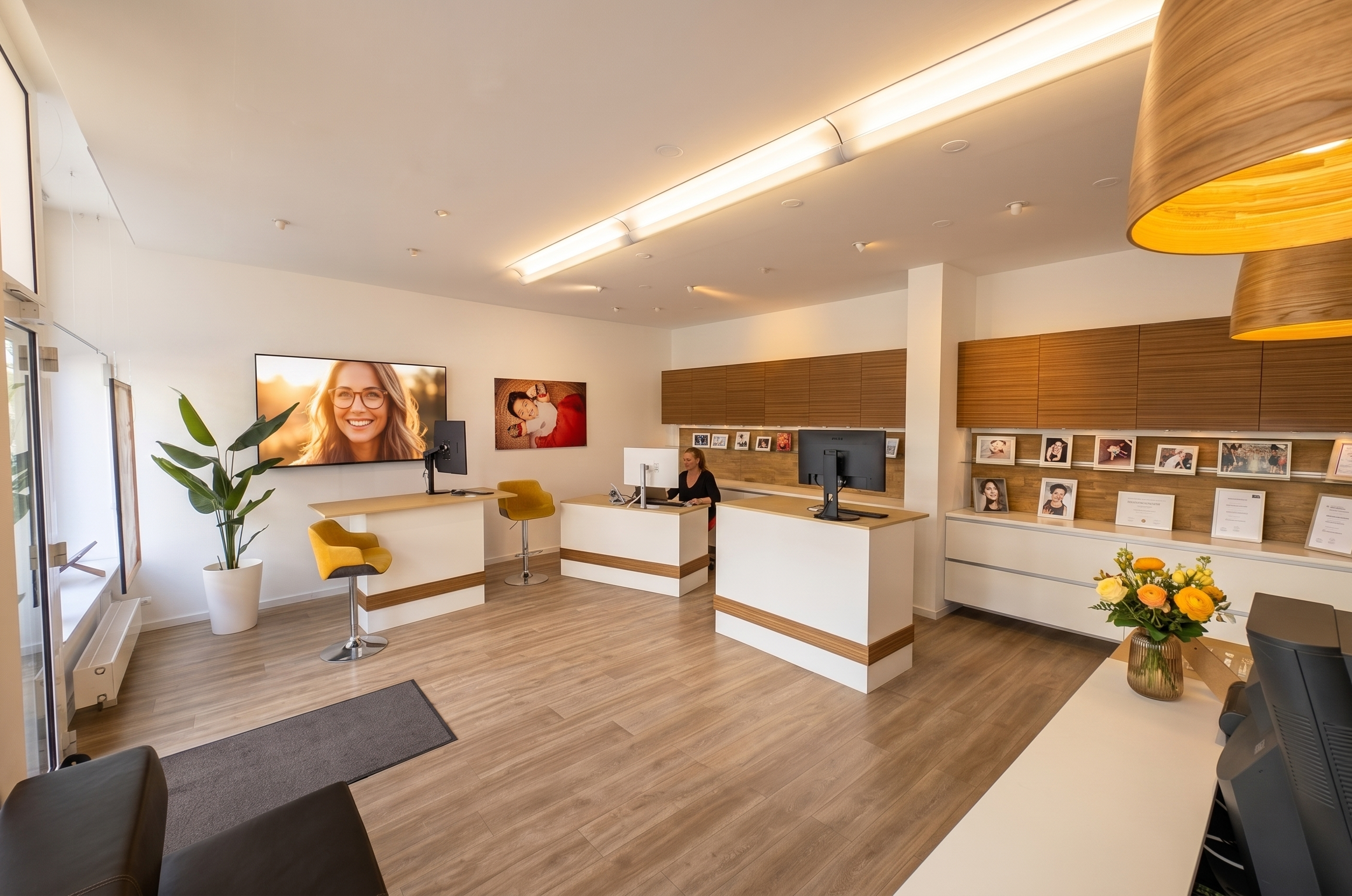 Modernes Fotostudio Hirch in Darmstadt mit Empfangstresen, einem Bildschirm mit einem Lächelnden Frauenbild, Farbige Fotos an der Wand, einem Gelb-weißen Sessel, Pflanzen und einem Blumenstrauß.