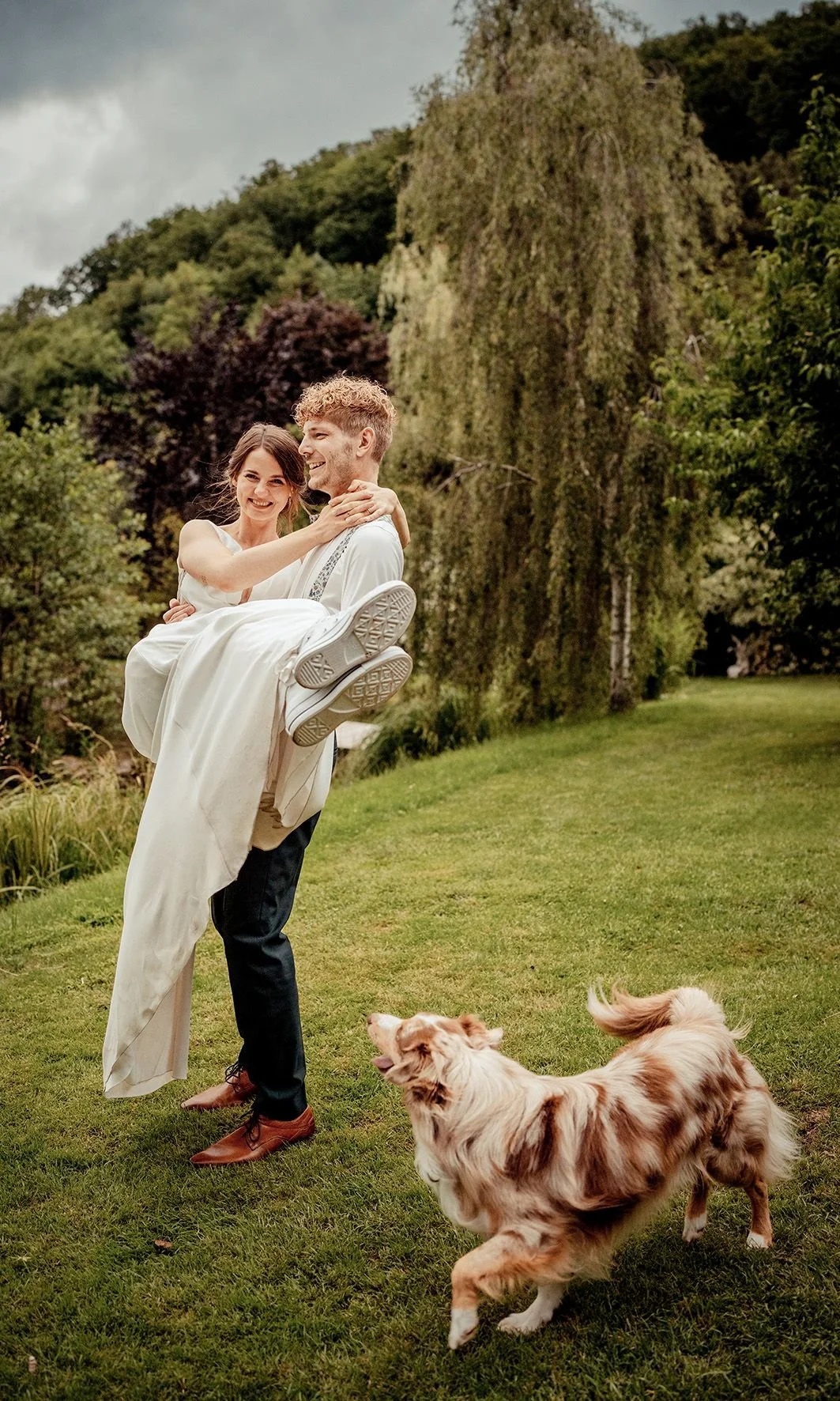 Ein Hochzeitspaar, der Mann trägt seine Frau. Im Vordergrund läuft ein Hund auf dem Rasen.
