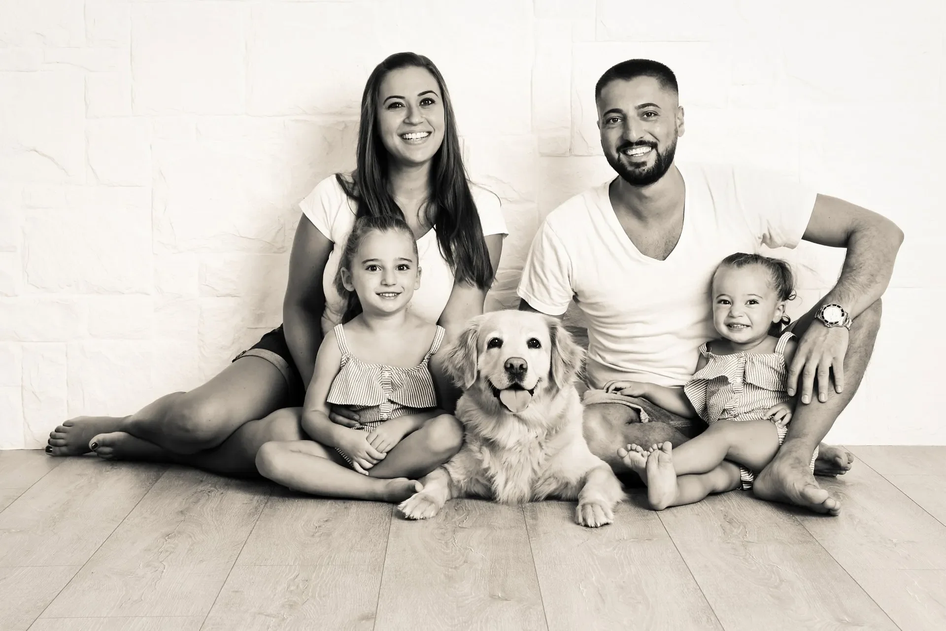 Familien Portrait Fotoshooting im Fotostudio in Darmstadt Eine Familie mit zwei kleinen Mädchen und einem Hund sitzt auf dem Boden vor einer weißen Wand, alle lächeln fröhlich in die Kamera.