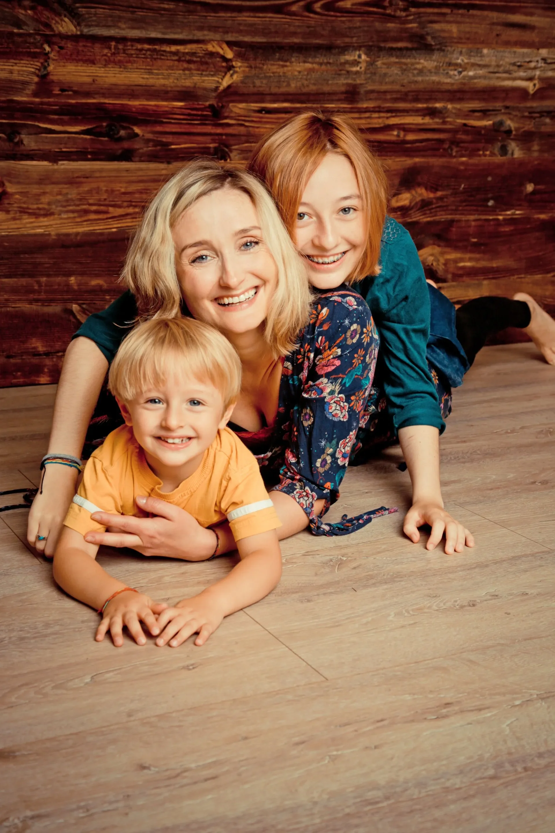 Familien Foto Shooting Eine Frau und zwei Kinder, liegen lachend auf einem Holzboden vor einer Holz-Wand, posieren für die Kamera.