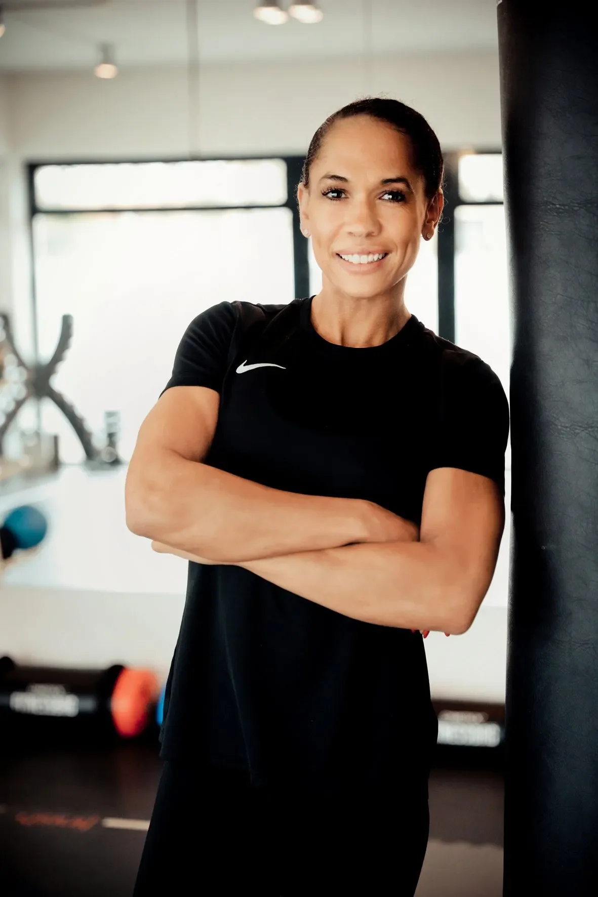 Fitnesstrainerin in Sportkleidung in Fitnessstudio, lächelnd, mit verschränkten Armen, vor einem Boxsack.