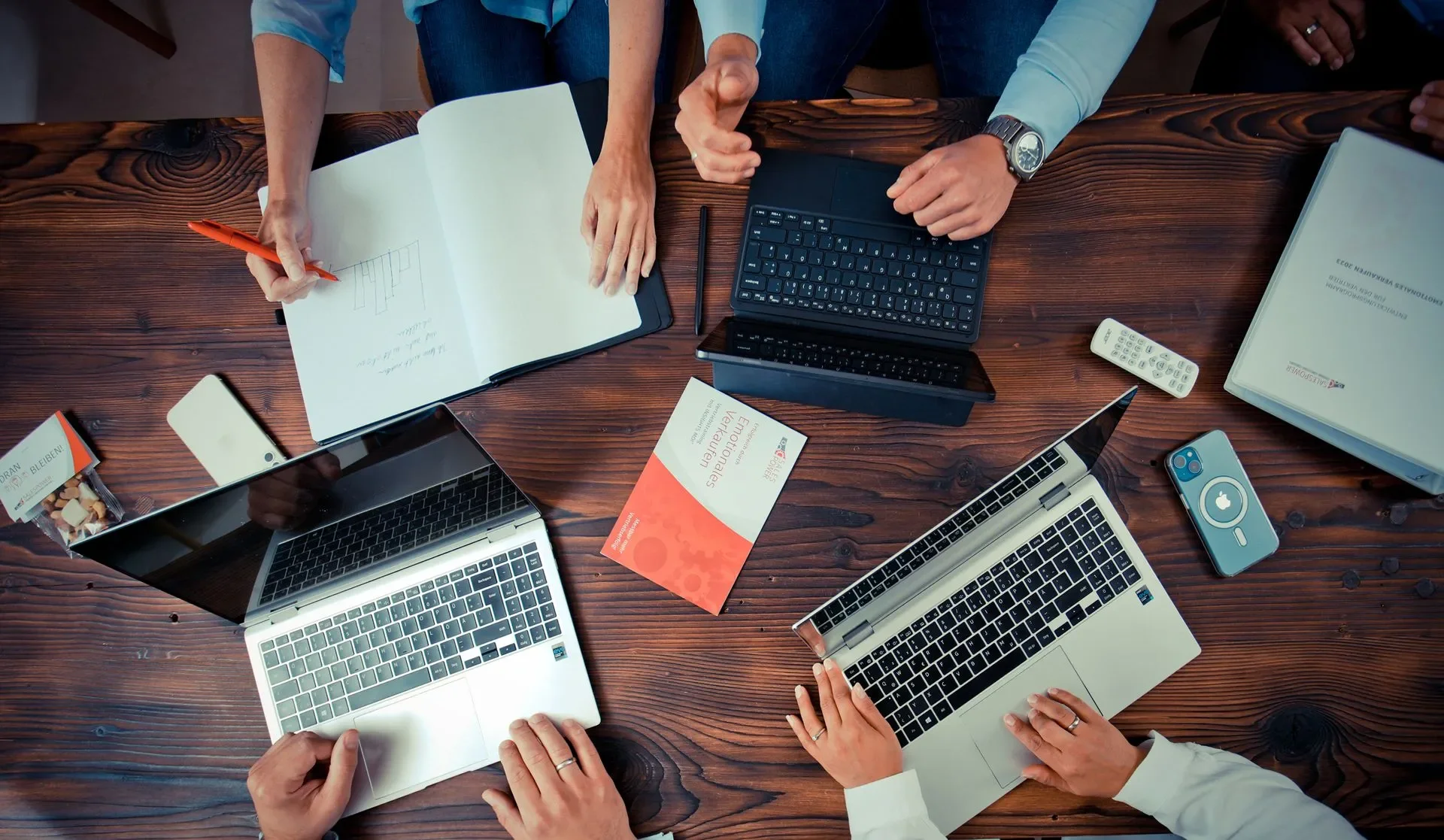 Überblick eines höhenverstellbaren Tisches mit mehreren Laptops, Handy, Dokumenten, Pens und anderen Büromaterialien, auf denen vier Menschen mit unterschiedlichen Händen arbeiten.