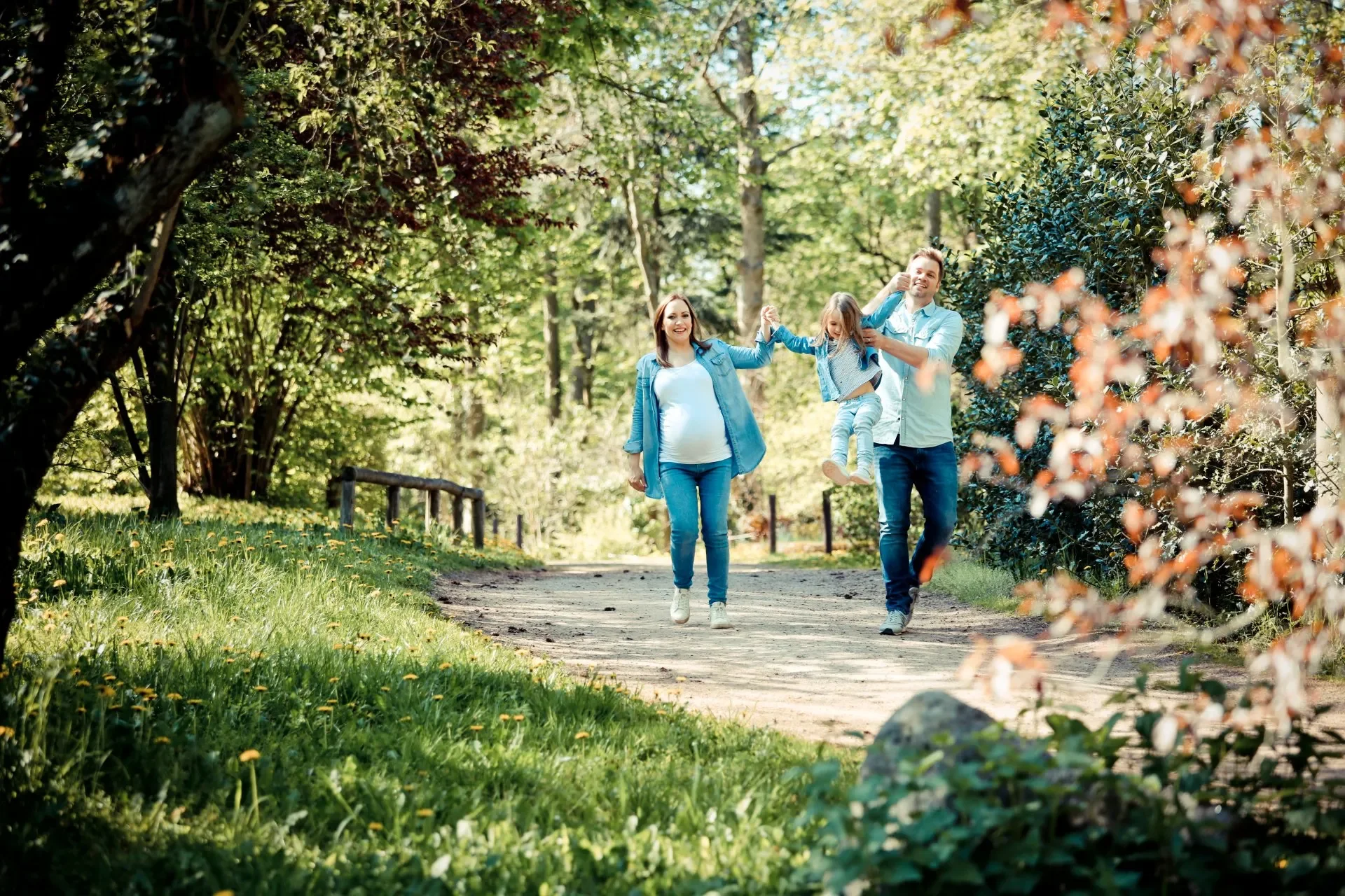 Familie mit Kind beim Spaziergang im Park an sonnigem Tag, umgeben von grünen Bäumen und Blumen.