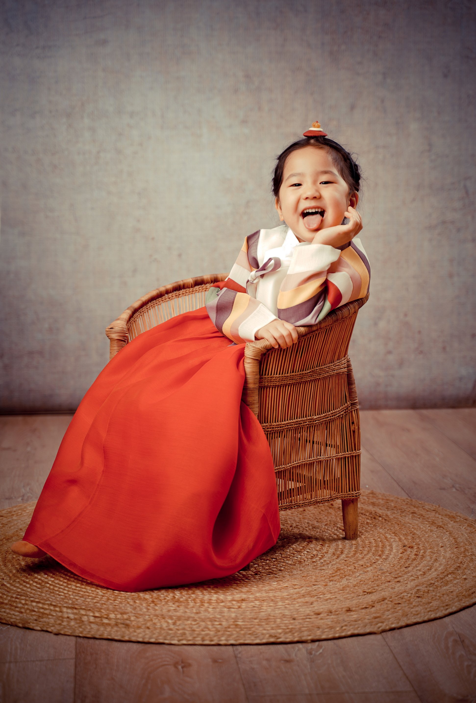 Ein lachendes Kleinkind in traditionellen koreanischen Hanboks, sitzend auf einem Korbstuhl, vor einem neutralen Hintergrund, mit einem runden Teppich darunter.