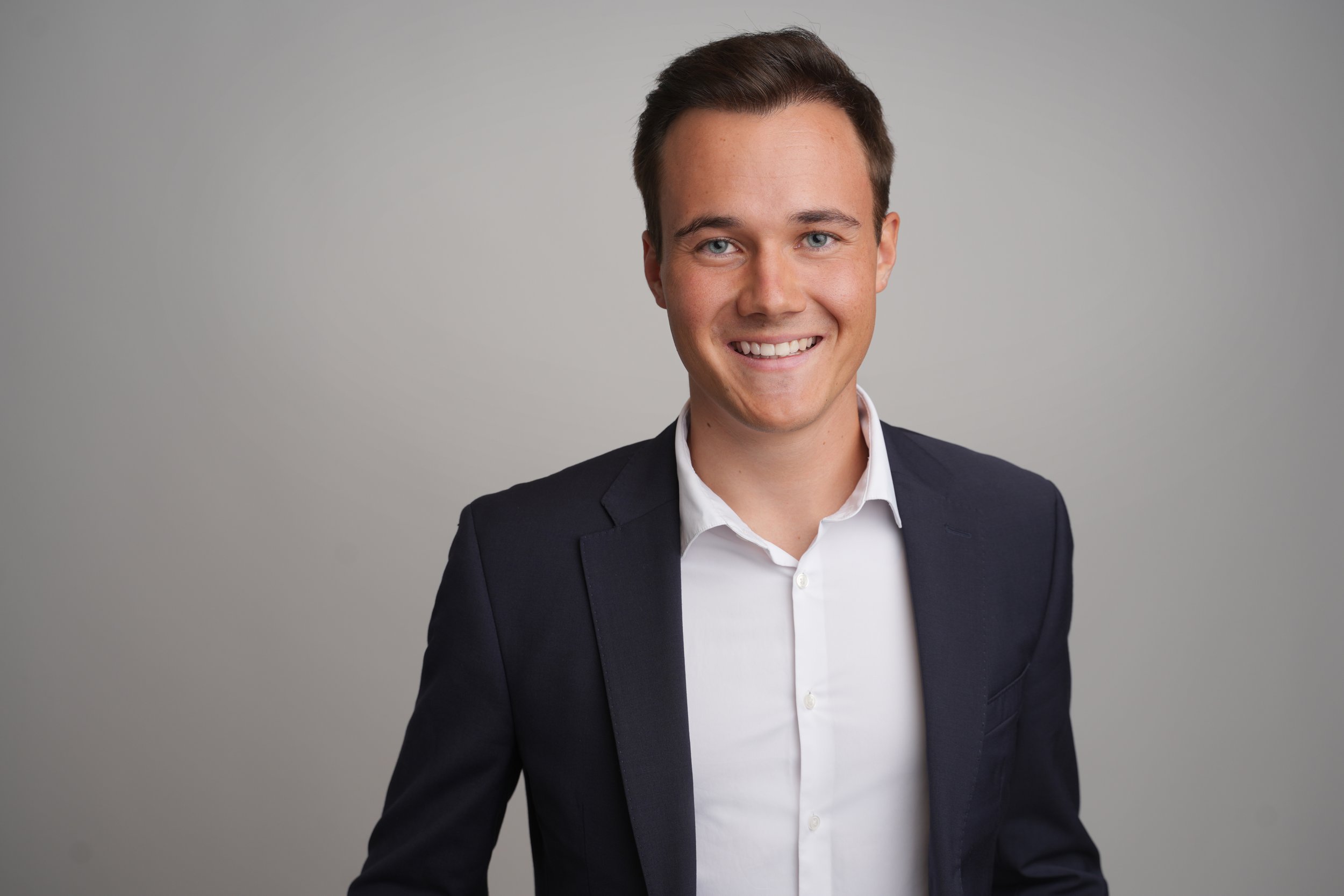 Modernes Bewerbungsfoto im Fotostudio in Darmstadt - Ein junger Mann trägt einen dunklen Anzug und ein weißes Hemd, lächelt und steht vor einem grauen Hintergrund.