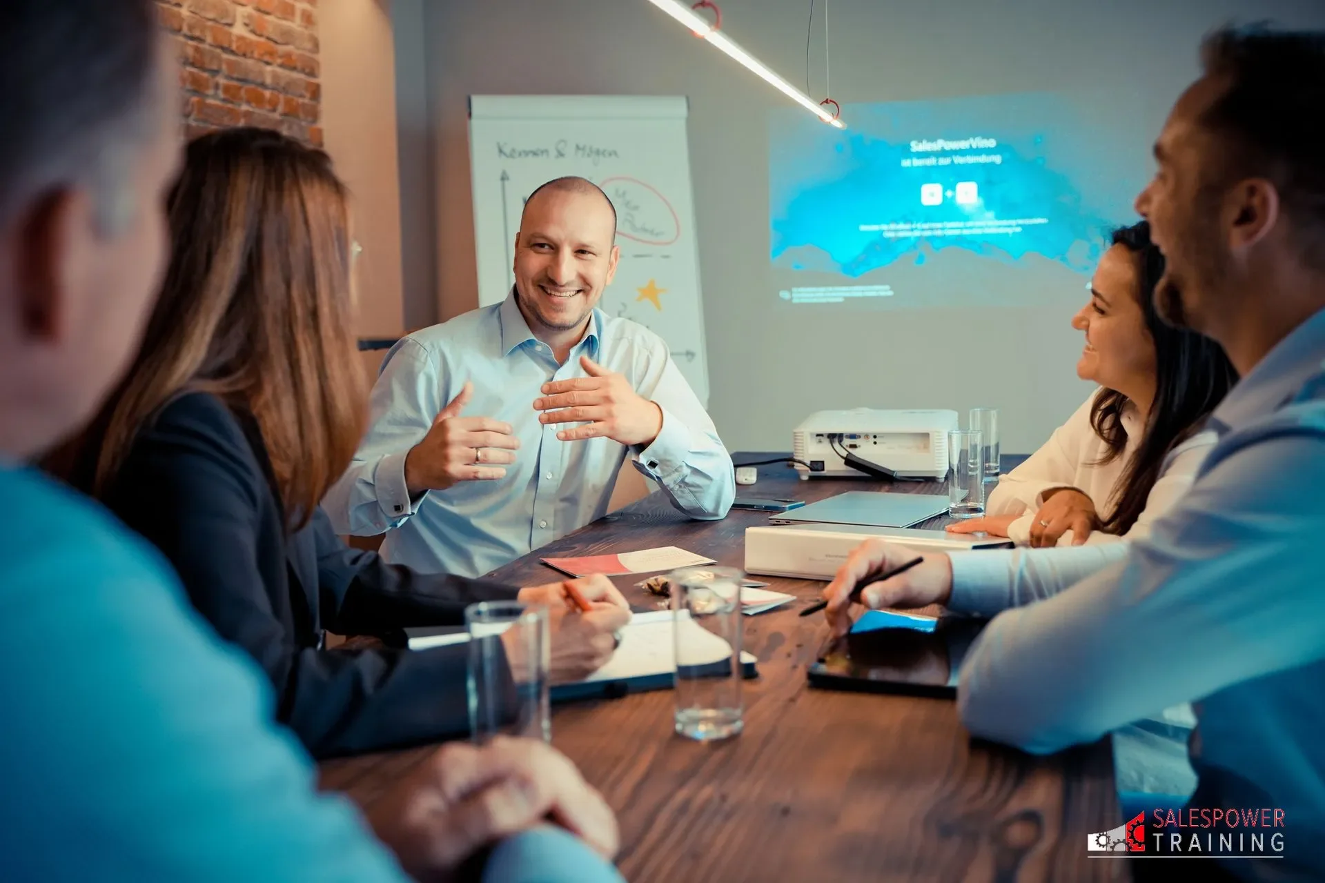 Eine Gruppe von Geschäftsleuten sitzt in einem Besprechungsraum um einen langen Tisch und führt eine Diskussion. Im Hintergrund ist ein Projektor, der eine Präsentation zeigt, und eine Whiteboard. Einer der Männer, der in der Mitte sitzt, spricht und