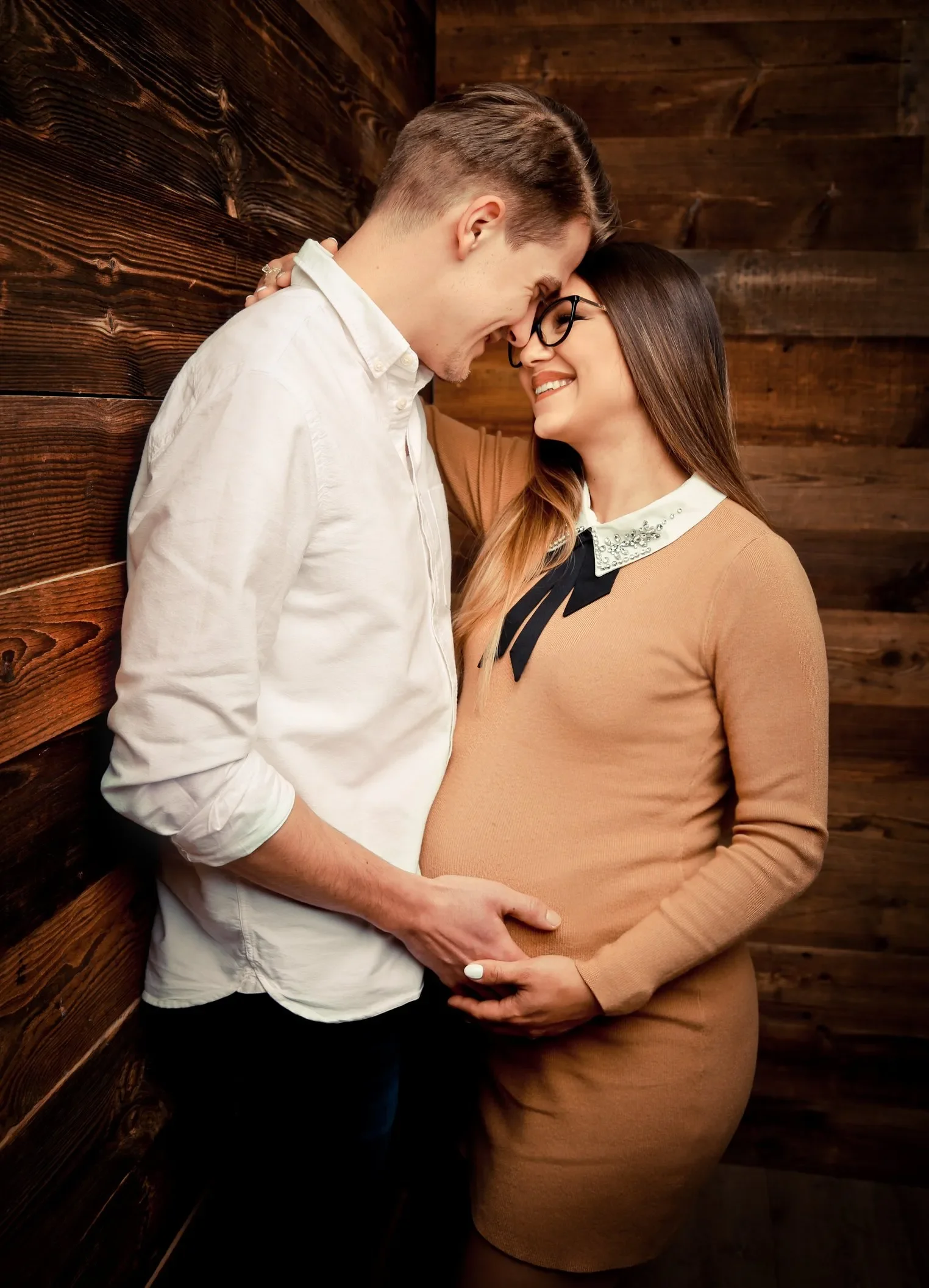 Babybauch Fotoshooting Bild. Ein verliebtes Paar, das seine Stirn und Nasen aneinanderlegt, während die schwangere Frau lächelt. Sie stehen vor einer Holz-Wand.