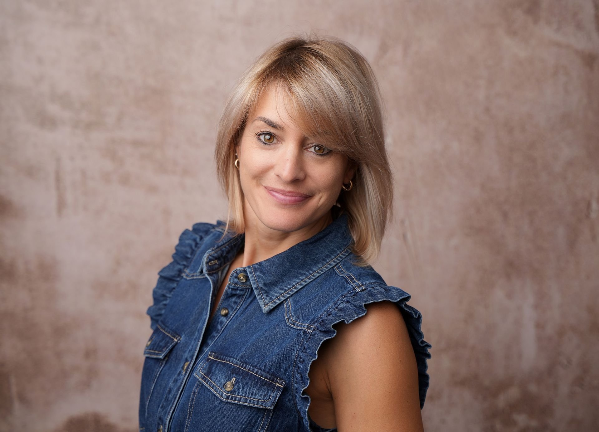 Junge Frau mit braunen, shoulder-langen Haaren, braunen Augen und lockeren Locken, trägt eine blaue Denim-Jacke mit Rüschen an den Schultern, lächelt und schaut in die Kamera vor einem neutralen beigen Hintergrund.