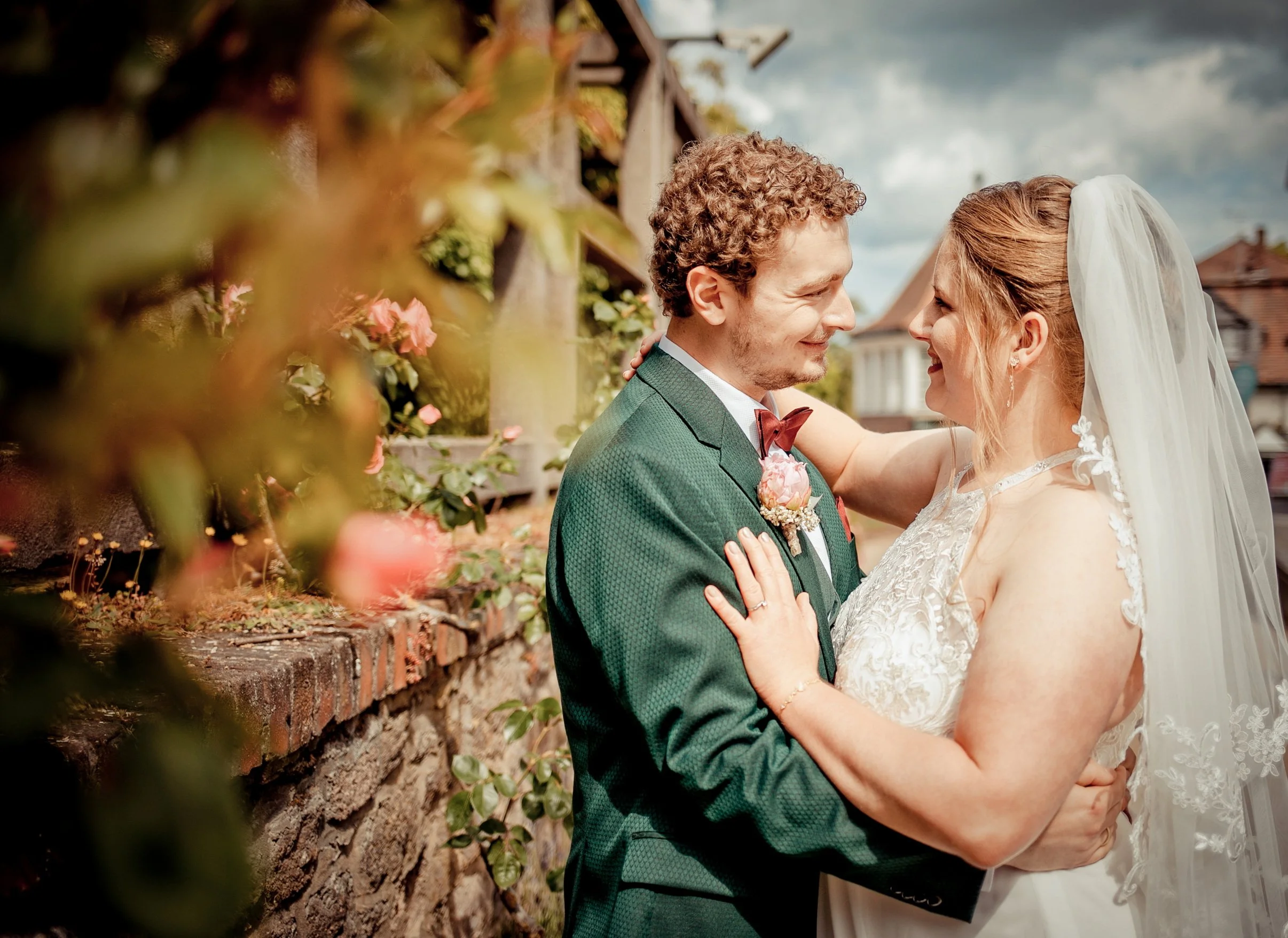 Ein Brautpaar beim Hochzeitstanz vor einer Ziegelmauer mit Blumen und einem bewölkten Himmel im Hintergrund.