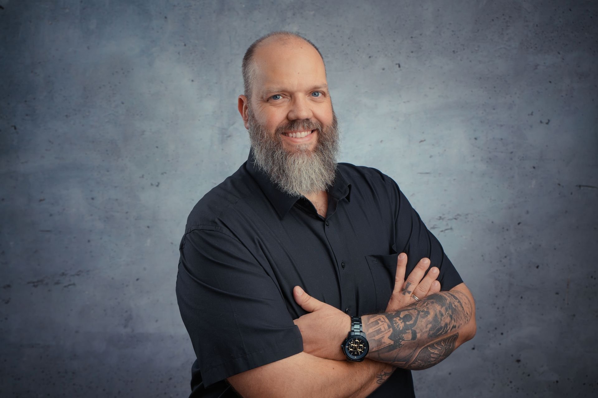 Bewerbungsfoto - Ein lächelnder Mann mit einem Bart und blauen Augen, trägt eine schwarze kurze Ärmelhemden, hat Tattoos am Arm, trägt eine schwarze Armbanduhr, steht vor einer grafitgrauen Wand, mit verschränkten Armen.