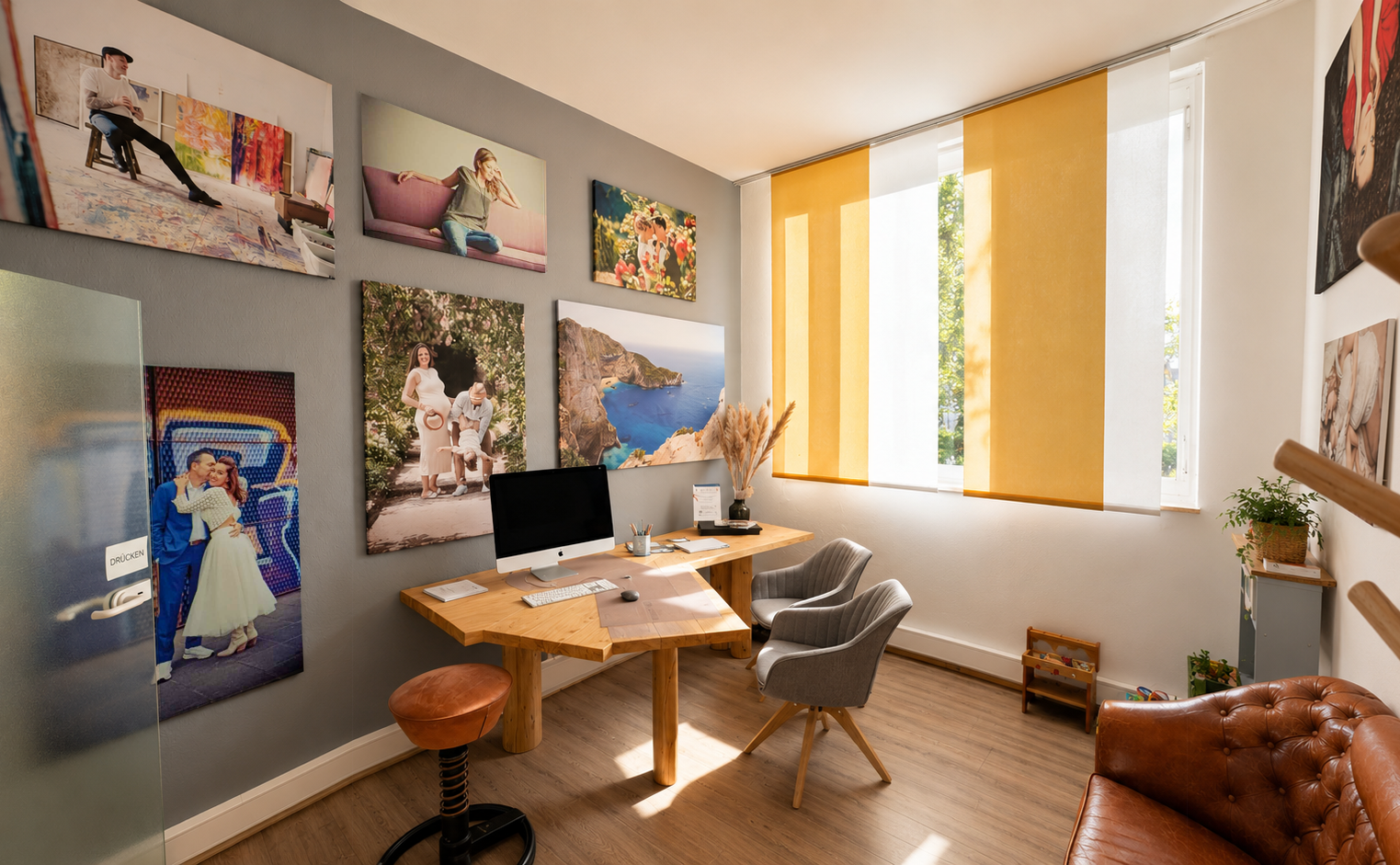 Büro mit einem Holzschreibtisch, zwei grauen Sessel, Wandkunst mit Fotos einer Frau und Landschaften, gelbe Vorhänge vor Fenstern, Pflanzen, Kinder Spielzeug in der Ecke.