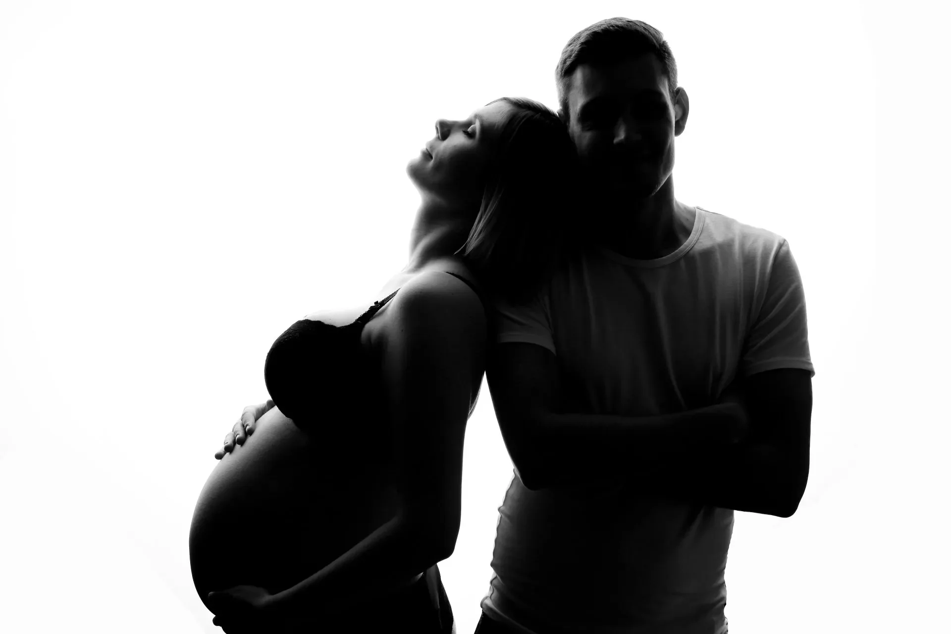 Silhouette eines Paares, eine Frau, die ihren Babybauch hält, und ein Mann, der sie umarmt, vor hellem Hintergrund, Schwarz-Weiß-Ästhetik.