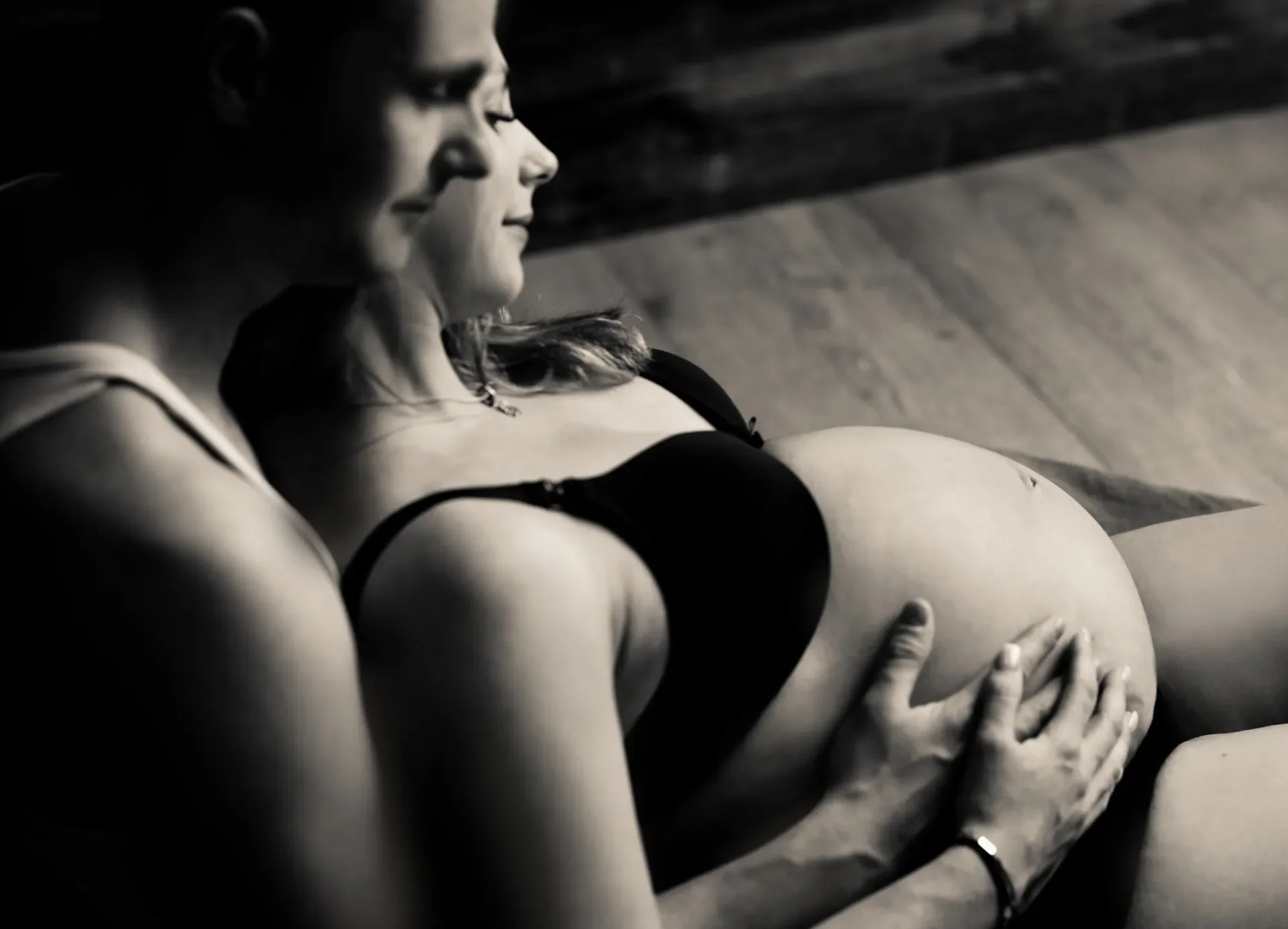 Babybauch Foto Shooting Schwarz-Weiß-Fotografie einer sitzenden Frau in einem Bikini, die auf dem Boden liegt, mit Blick nach unten. Im Hintergrund Holzdielenboden.