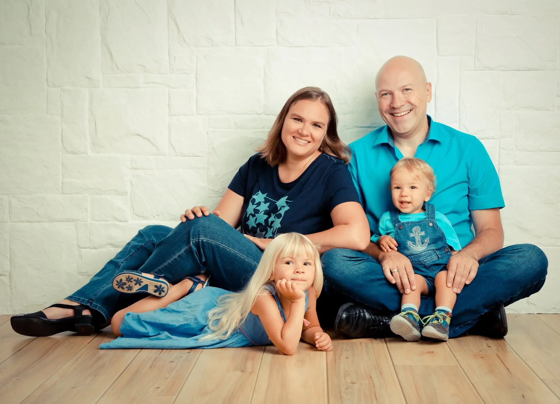 Familien Fotoshooting im Fotostudio. Eine Familie mit zwei erwachsenen Erwachsenen und drei Kindern sitzt auf einem Holzboden vor einer weißen Ziegelwand, lächelnd für ein Foto.