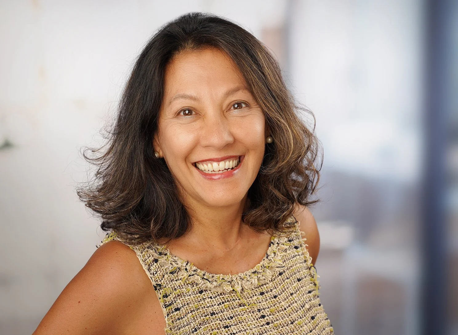 Business Portrait einer lachenden Frau mit dunklen, gewellten Haaren vor einem verschwommenen Hintergrund.