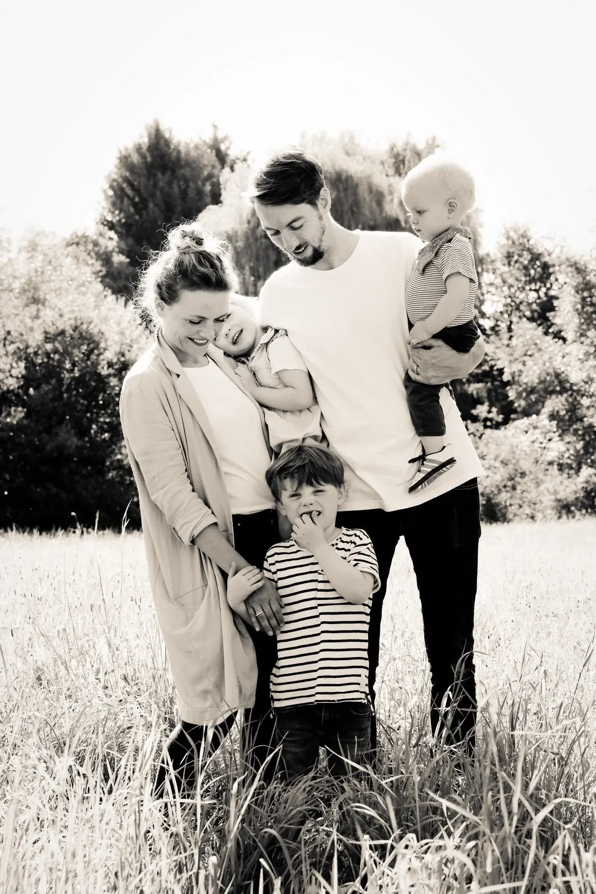 Familie mit zwei Erwachsenen und drei Kindern beim Spaziergang im Freien, lachend und umarmend, in einer Wiese mit Bäumen im Hintergrund, schwarz-weiß.