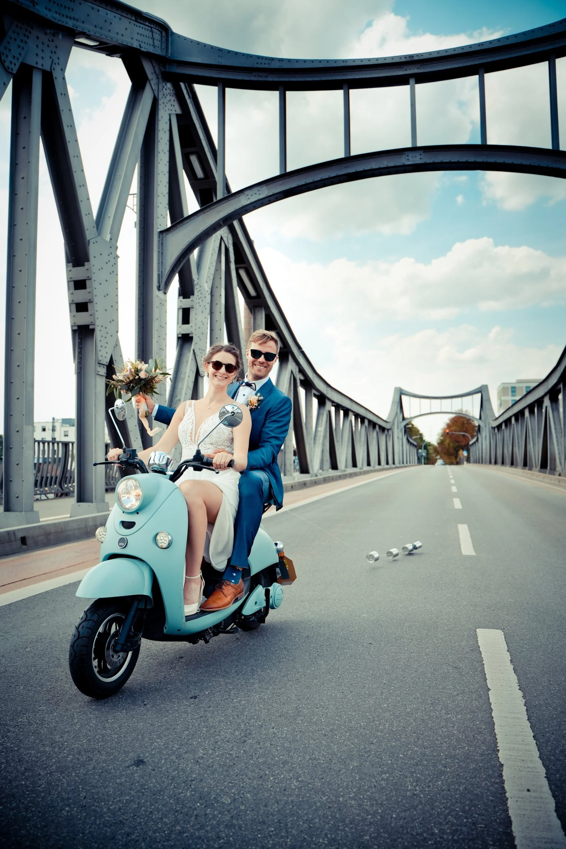 Ein Hochzeitspaar, das auf einem blauen Motorroller auf einer Brücke fährt, mit Wolken am Himmel.