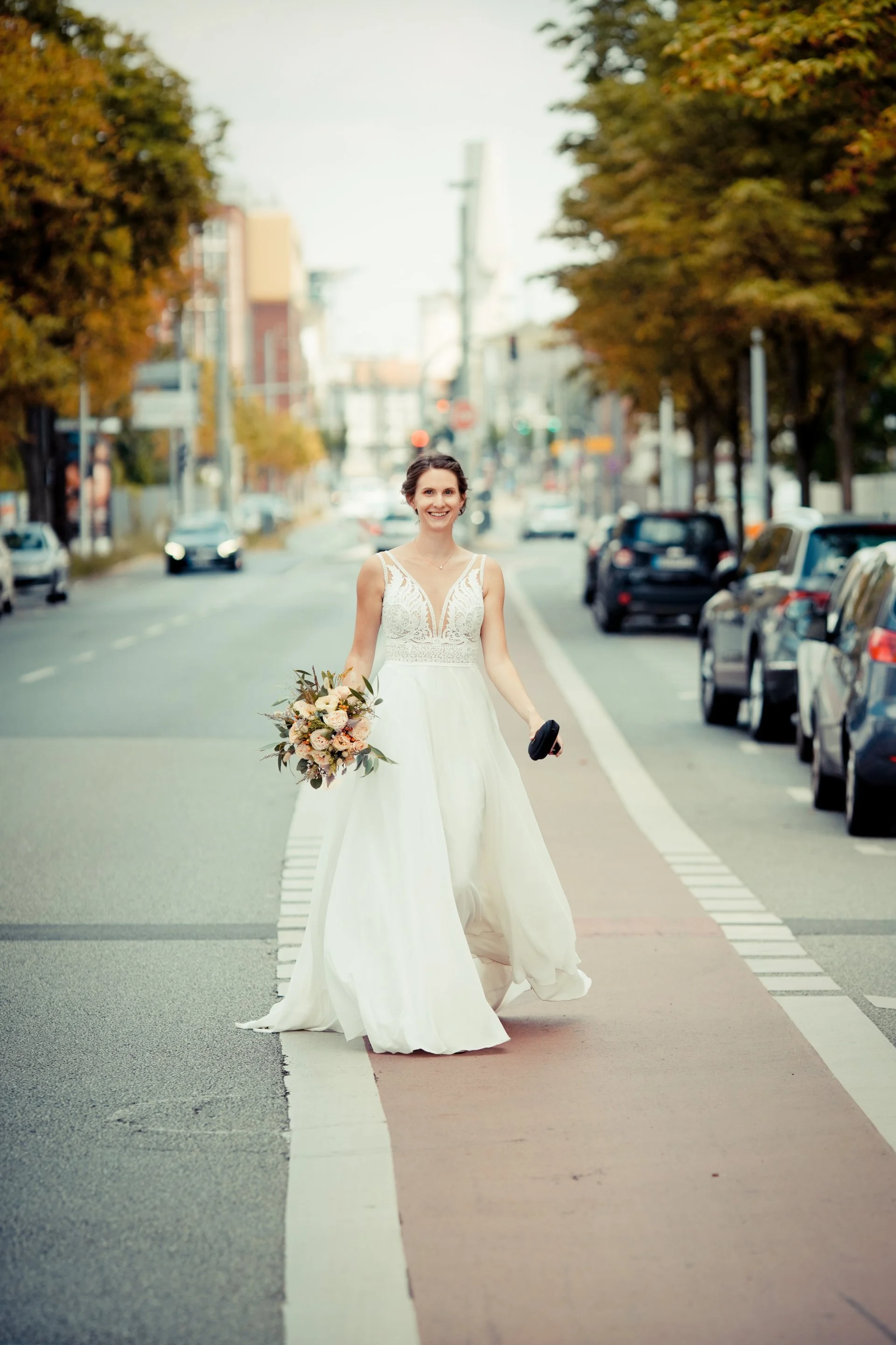 Eine lächelnde Frau in einem weißen Hochzeitskleid hält einen Blumenstrauß und geht auf einer Stadtstraße. Im Hintergrund sind Autos und Bäume mit Herbstlaub.