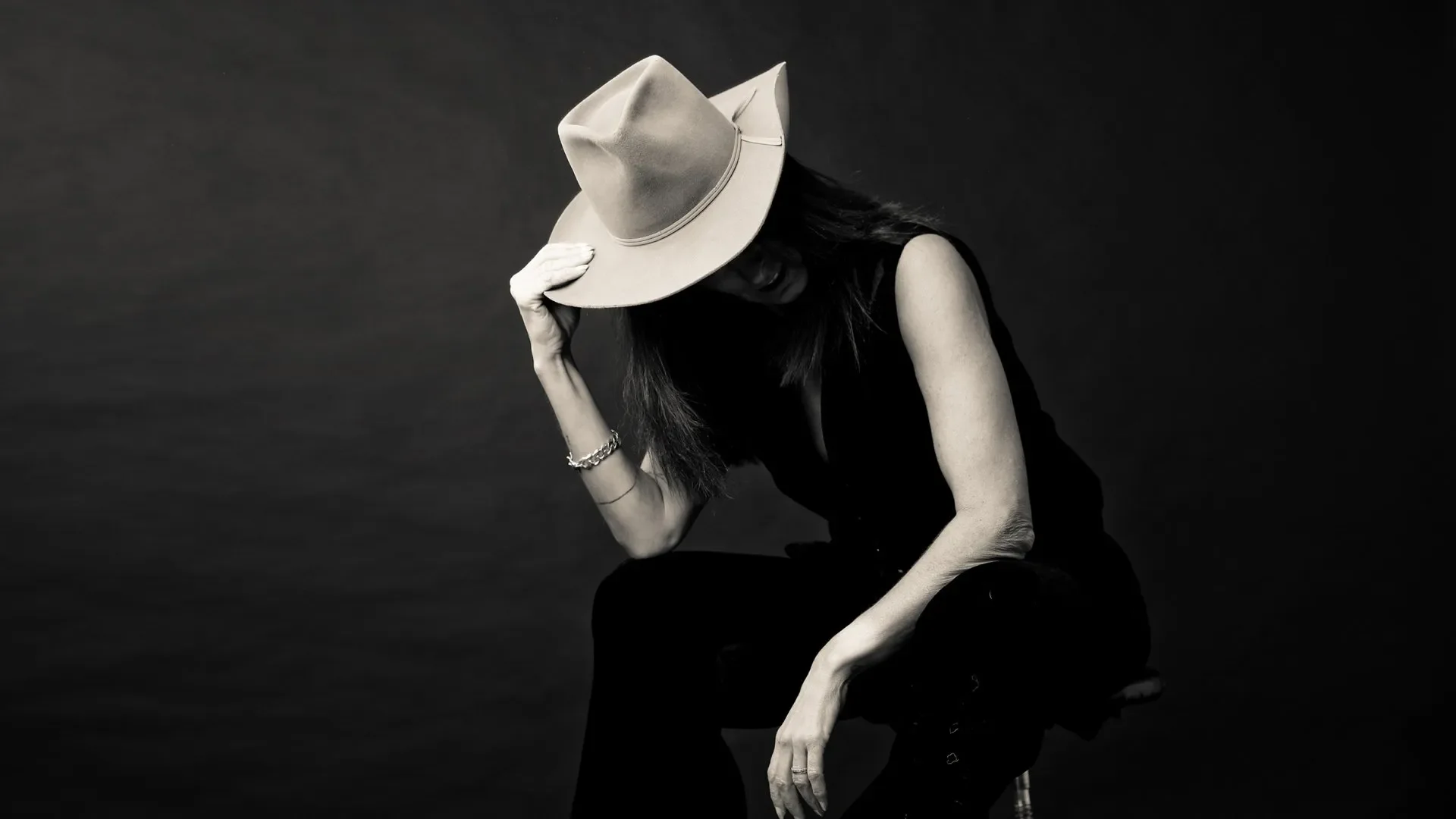Portrait Aufnahme - Frau trägt einen Cowboyhut und schwarze Kleidung, sitzt nachdenklich auf einem Stuhl, die Hand am Hut, vor dunklem Hintergrund.