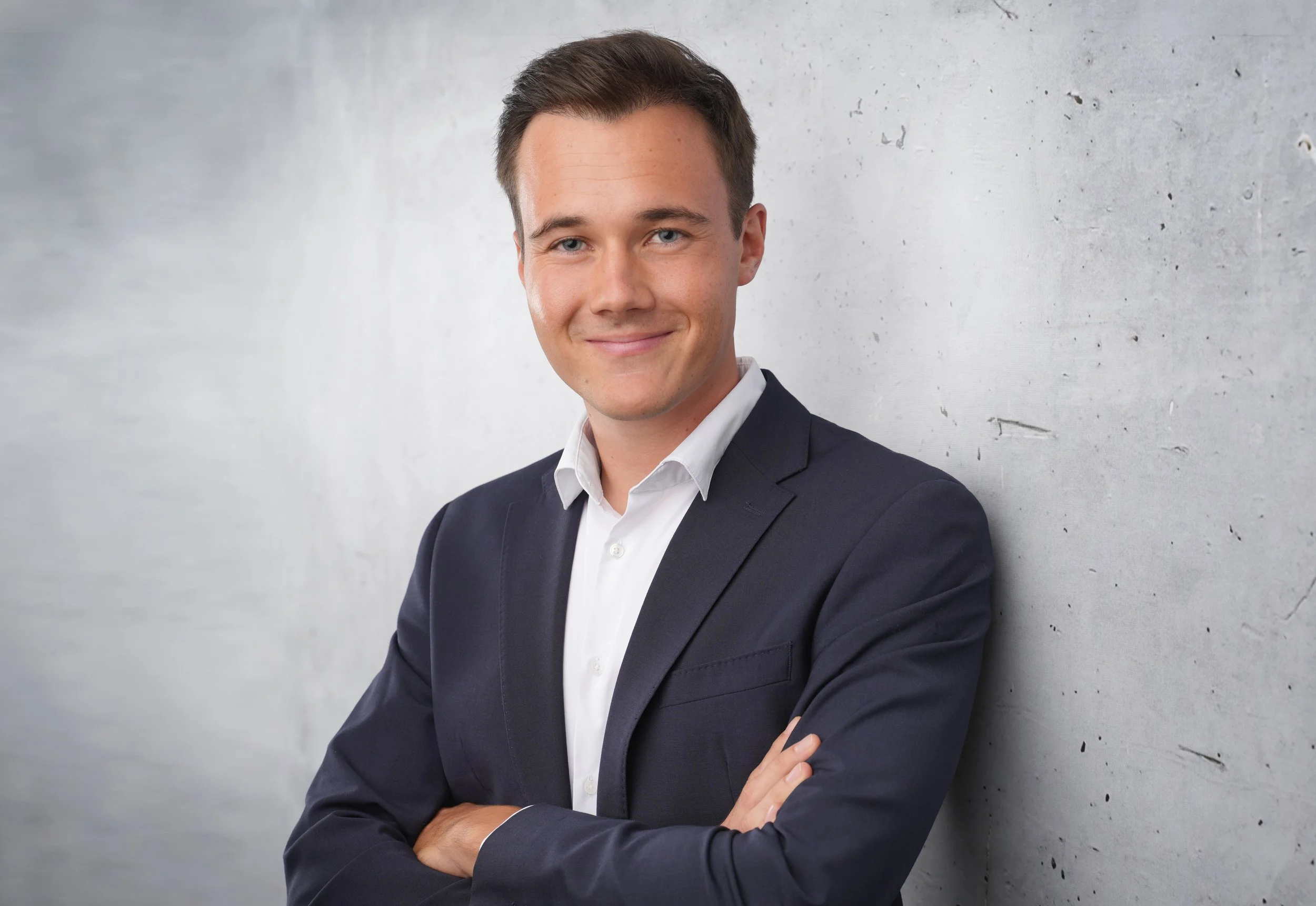 Bewerbungsbild von einem Mann in dunklem Anzug steht mit verschränkten Armen vor einer Betonwand und lächelt.