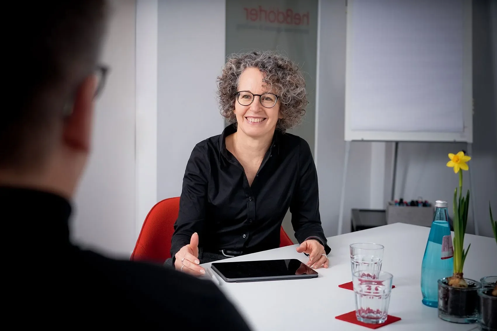 Eine Frau mit lockigem Haar und Brille sitzt an einem Tisch und spricht mit einem Mann, der nur teilweise im Bild sichtbar ist.