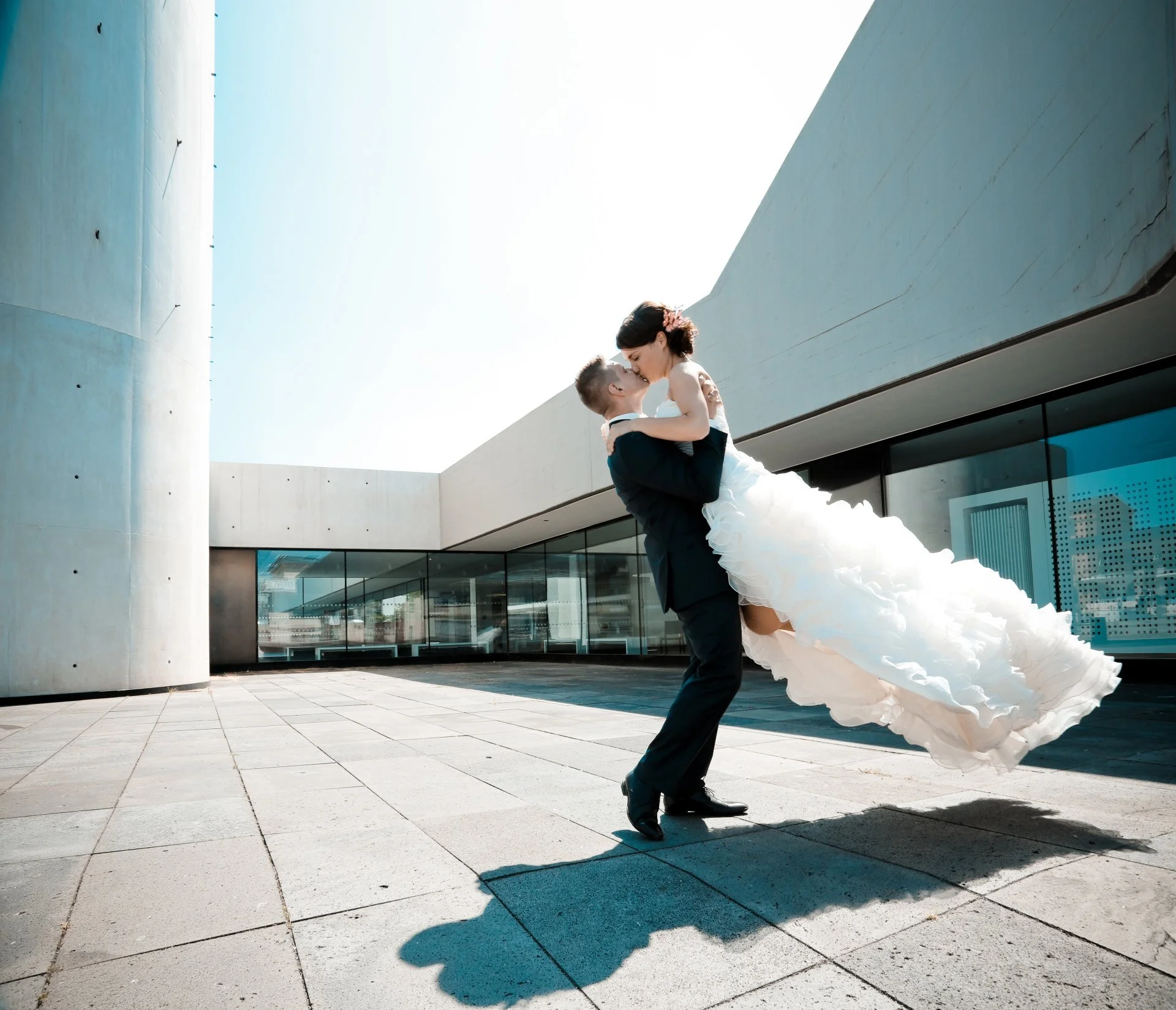Ein Mann in einem Anzug hält eine Frau in einem weißen Brautkleid hoch, während sie sich küssen. Sie befinden sich im Freien vor einer modernen, weißen Architektur mit Glasfenstern, bei sonnigem Himmel.