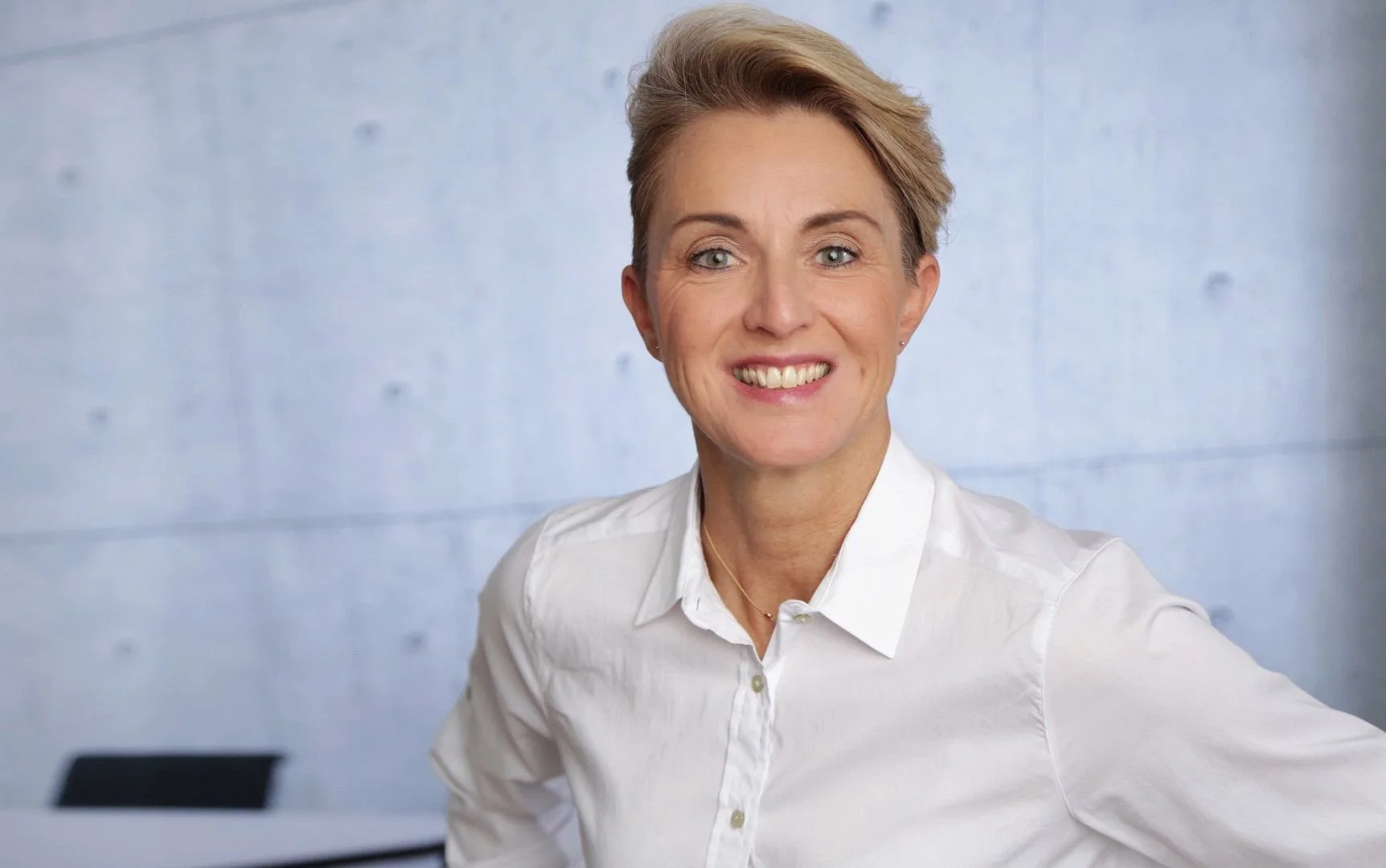 Business Portrait - Eine lächelnde Frau mit kurzen, seitlich stylierten, hellbraunen Haaren trägt eine weiße Hemd und steht vor einer hellblauen Wand.