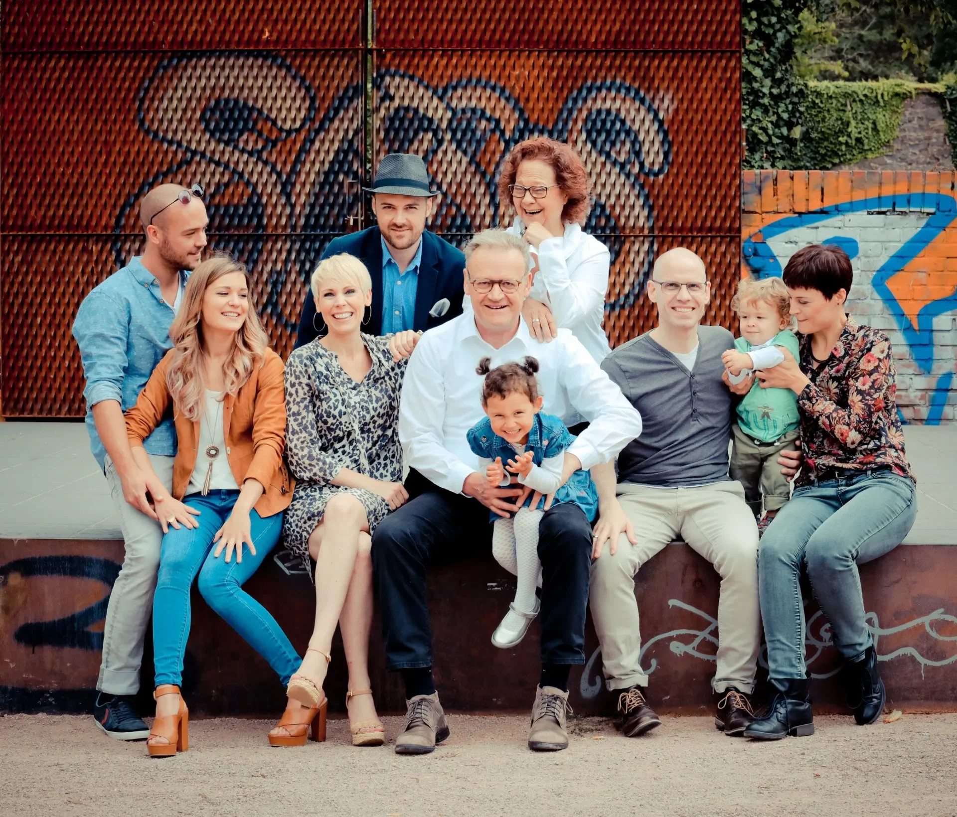 Gruppenfoto von Erwachsenen und Kindern, die zusammen auf einer Bank im Freien sitzen, vor einer Graffiti-verwenden Wand.
