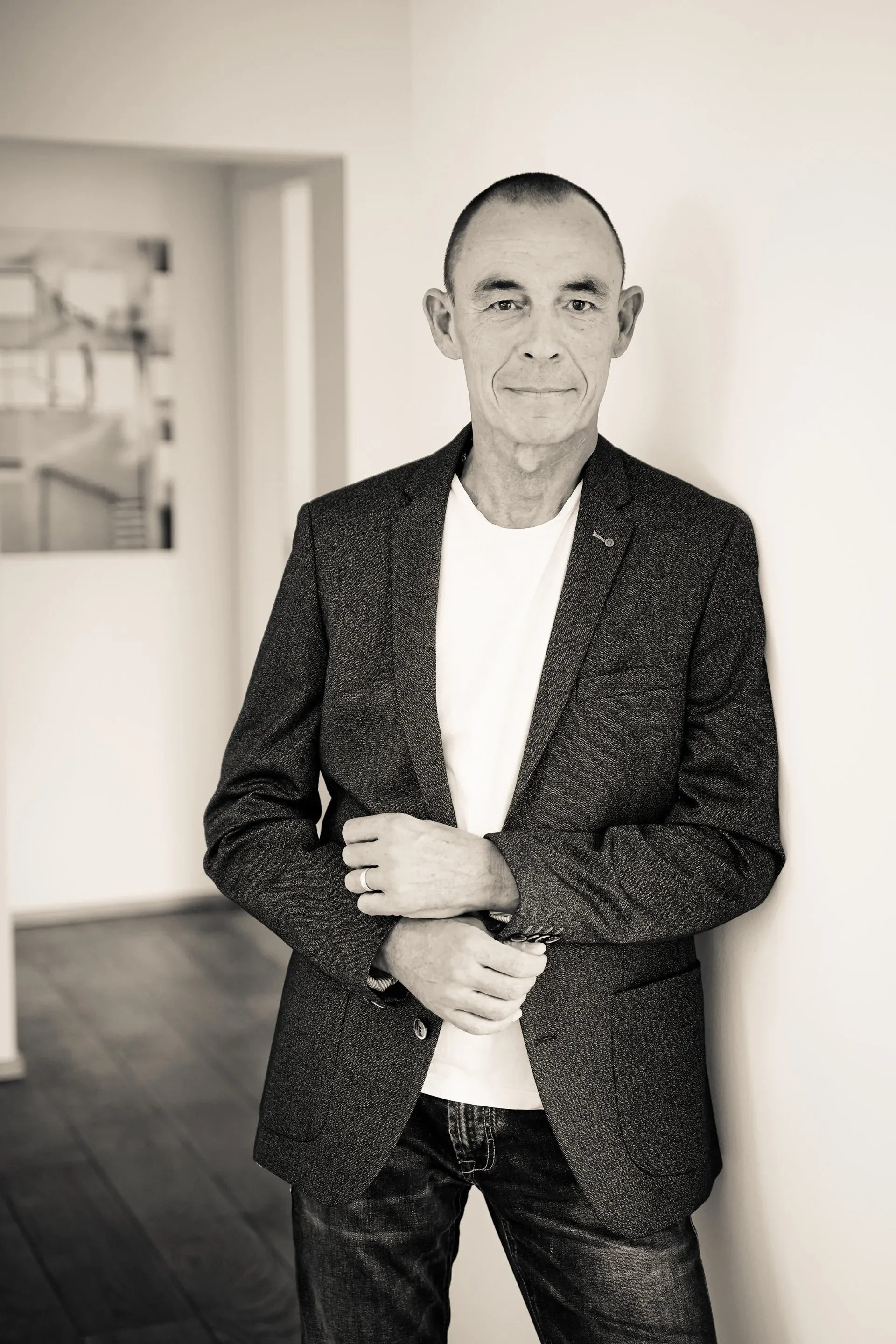 Business Portrait Schwarz-weiß-Foto eines Mannes mit kurzer Frisur, Anzugjacke und weißem T-Shirt, der gegen eine weiße Wand lehnt.