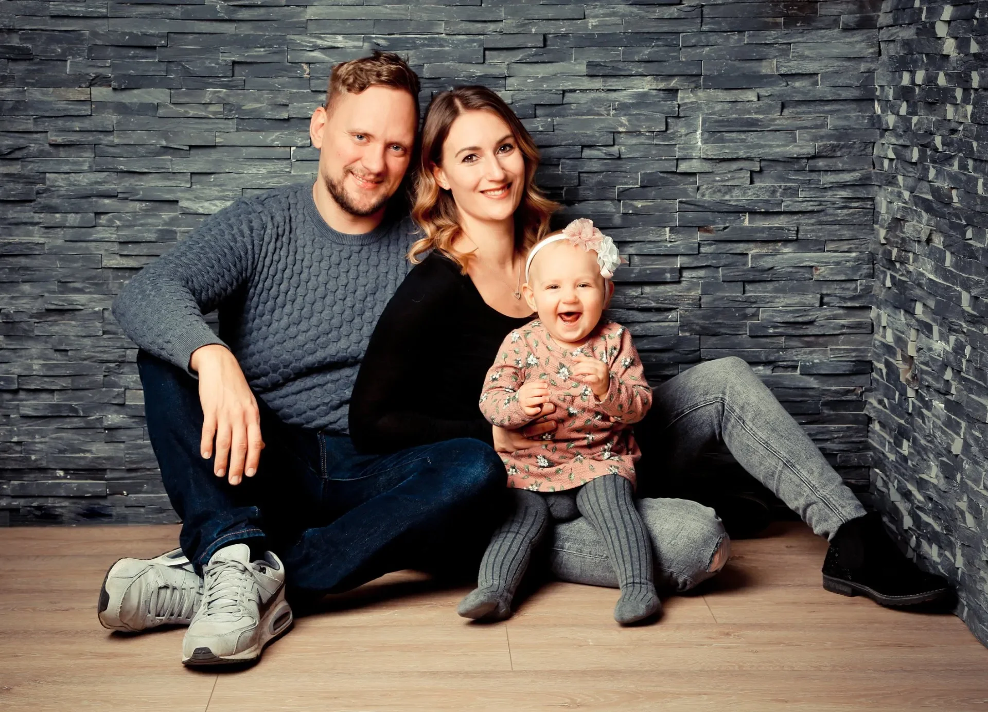Familien Portrait Fotoshooting - Familie sitzt lachend vor einer dunklen Steingwall hinter einem Holzboden, Vater, Mutter und kleines Mädchen, sie tragen lässige Kleidung und lächeln.