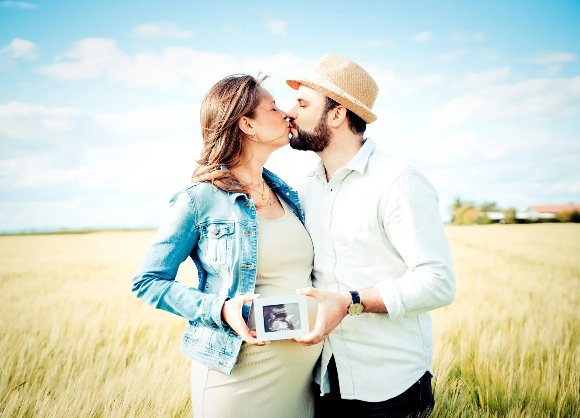 Babybauch Shooting Outdoor - Ein Paar küsst sich im Freien auf einer Weide, während die Frau mit einem Ultraschallbild in den Händen schwanger ist.