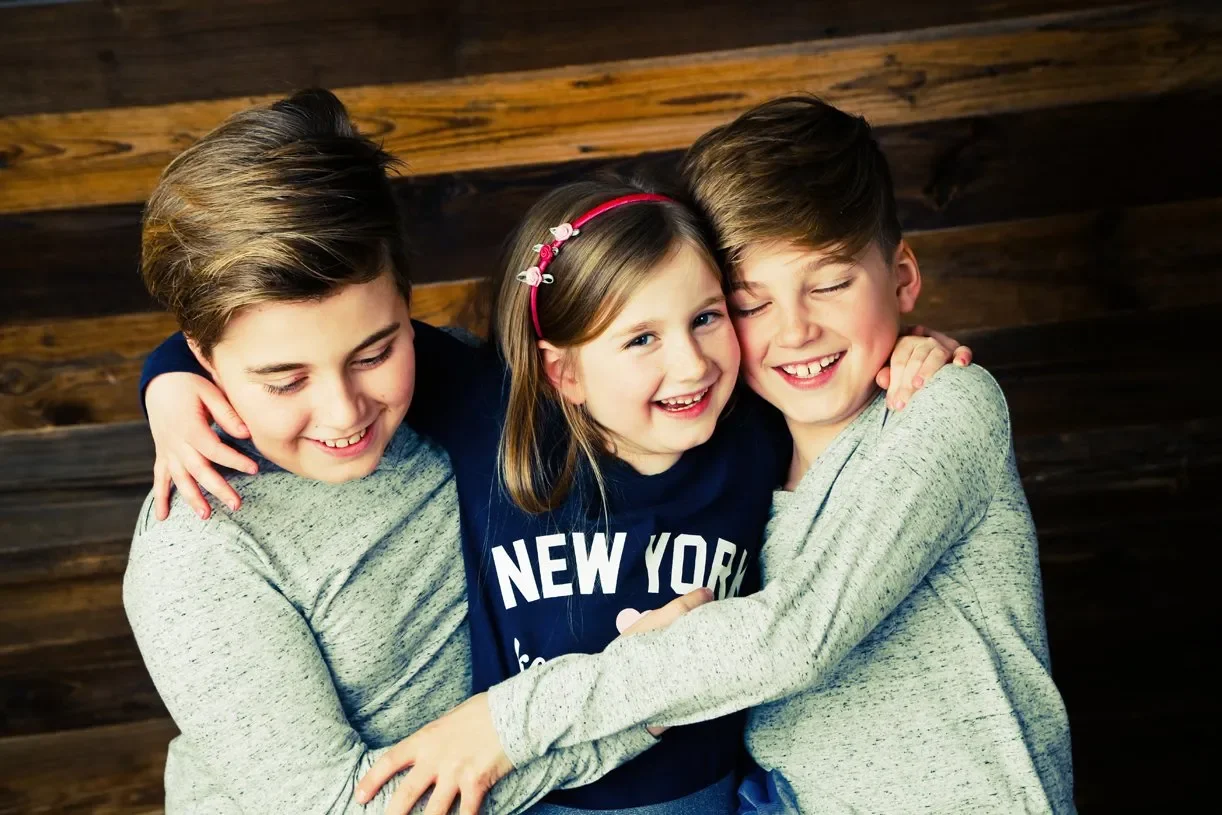 Geschwisterfotoshooting - Drei Kinder umarmen sich lachend vor einer Holzwand.