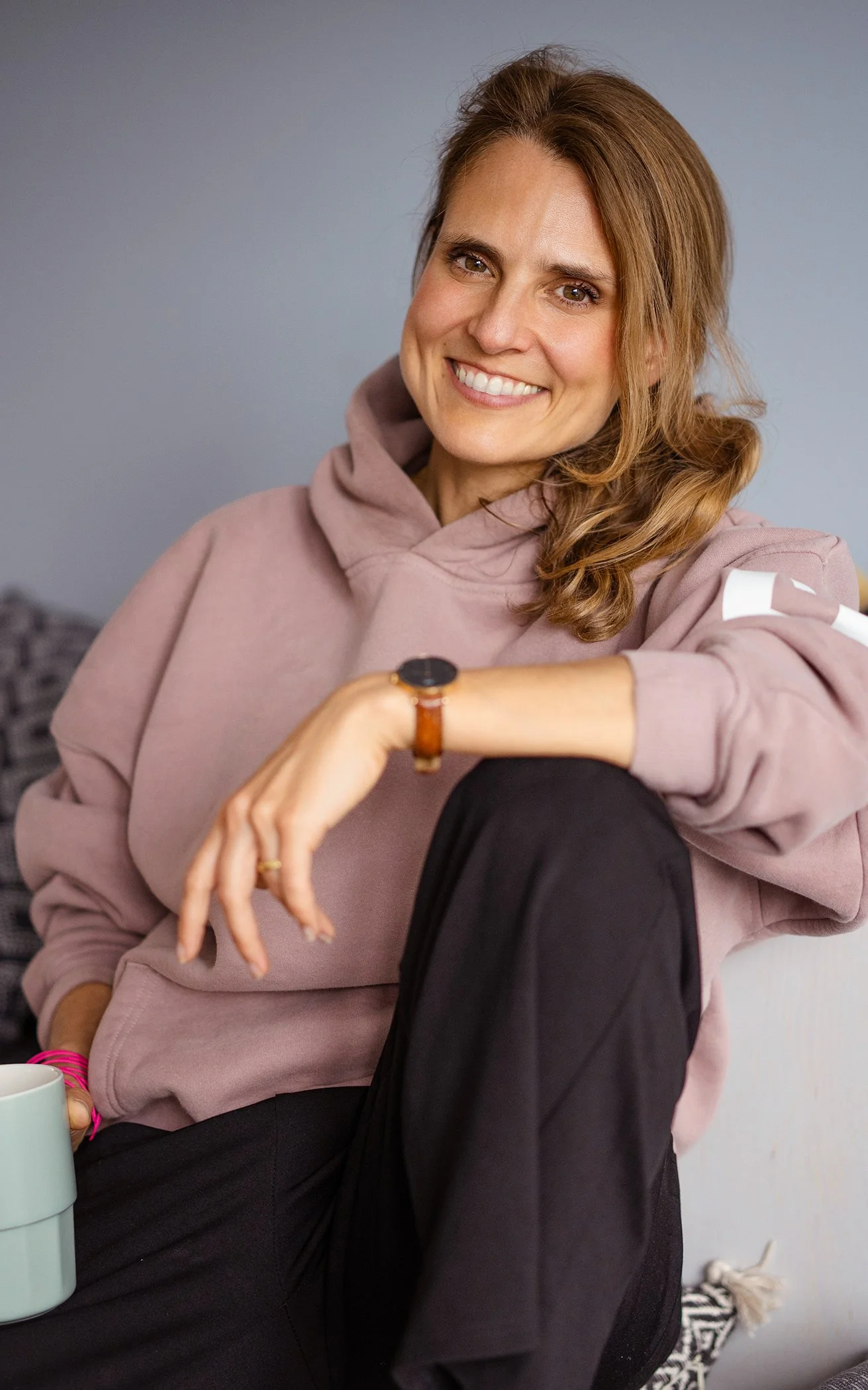 Lächelnde Frau mit braunen, gewellten Haaren trägt einen rosa Hoodie und sitzt entspannt im Raum.