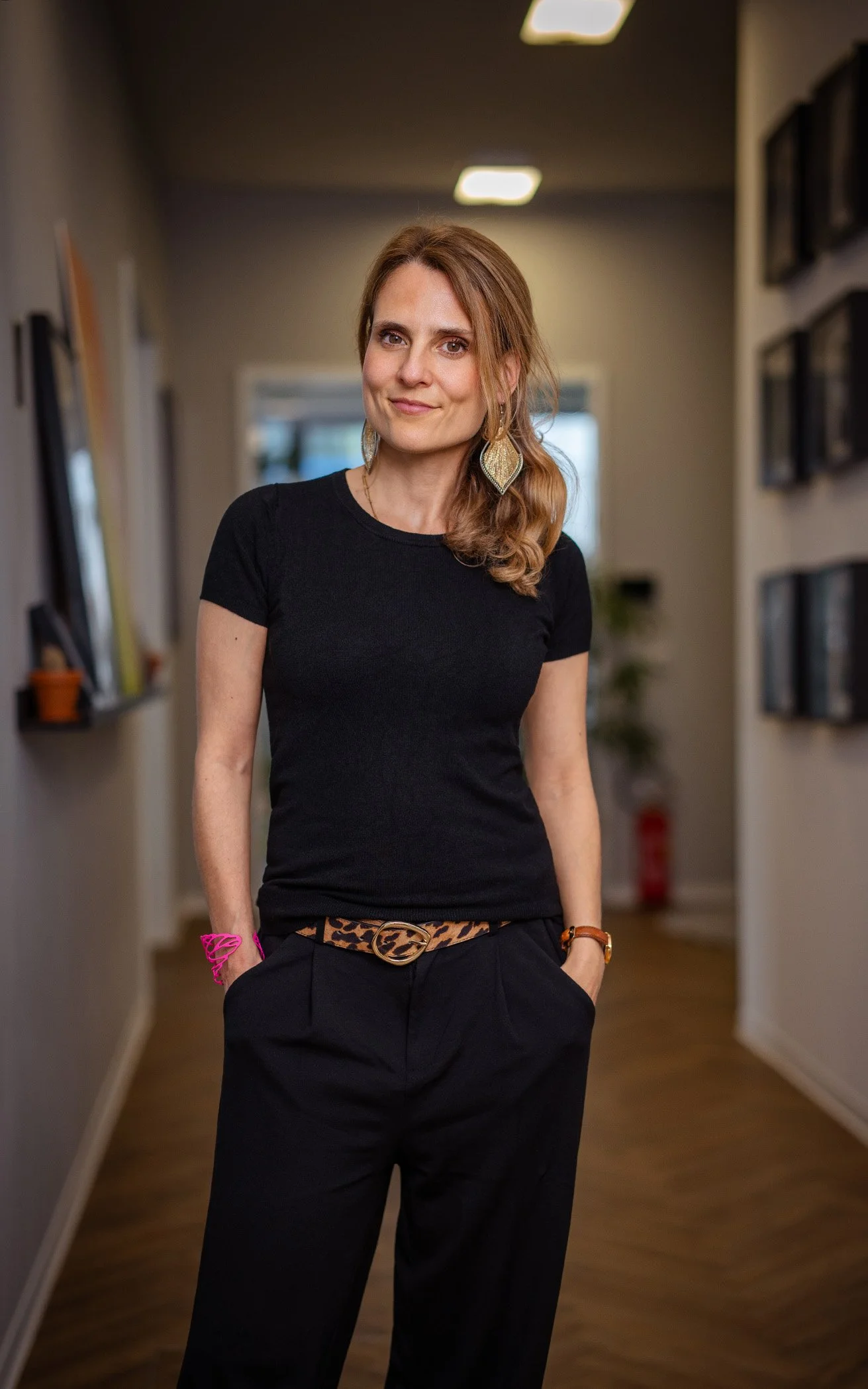Eine Frau steht in einem Flur mit dunklem Boden und gestrichenen Wänden. Sie trägt eine schwarze T-Shirt, eine Leoparden-Jeans und Accessoires wie Ohrringe, Armband und Gürtel. Im Hintergrund sind Bücherregale an den Wänden zu sehen.