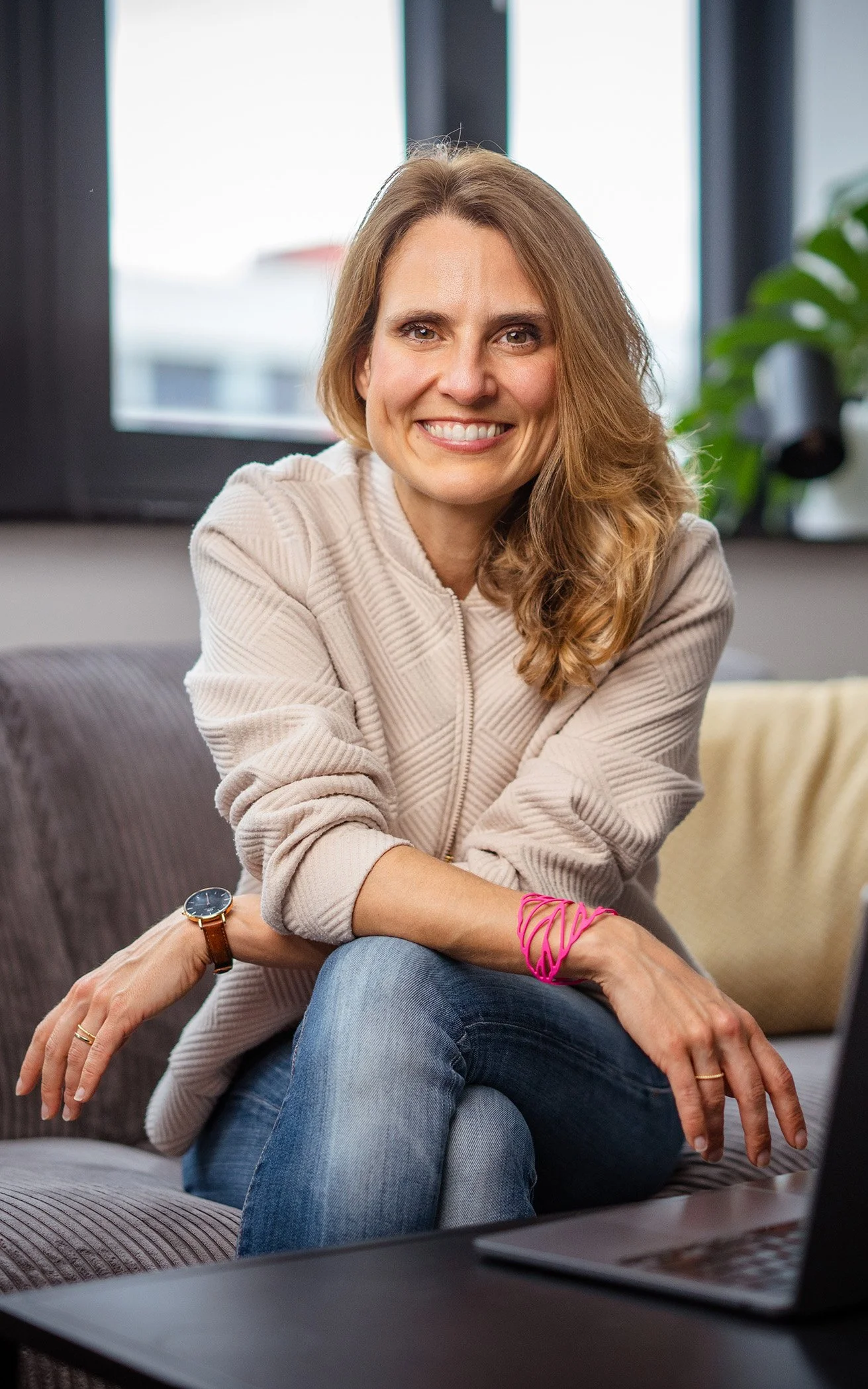 Eine lächelnde junge Frau mit braunen, welligen Haaren sitzt auf einem Sofa vor einem Fenster. Sie trägt eine beige Jacke, blaue Jeans, eine Armbanduhr und bunte Armbänder. Im Vordergrund ist ein Laptop auf einem Tisch sichtbar.
